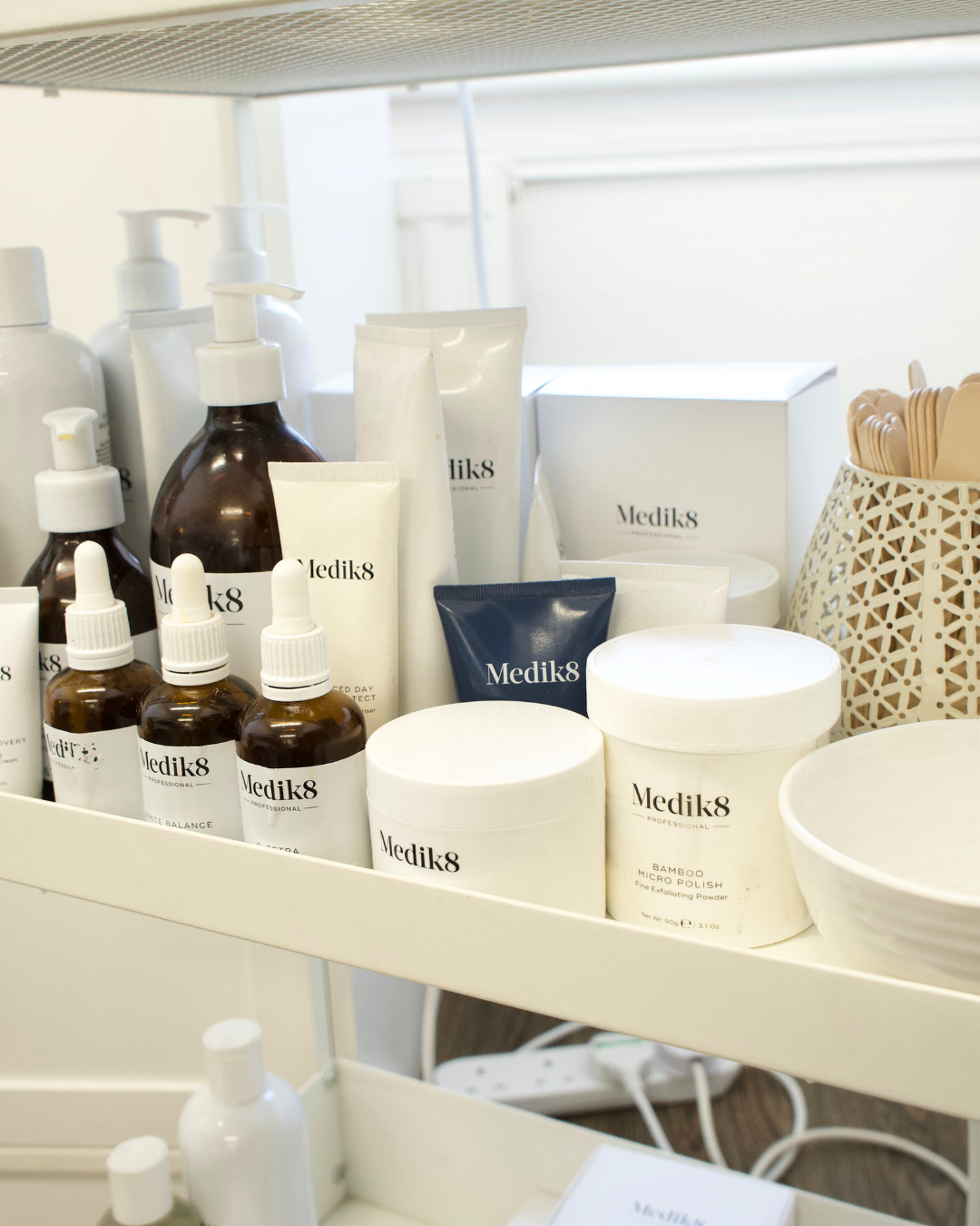 Collection of skincare and cosmetic products branded Medik8 on a white shelf, including bottles, jars, tubes, and a decorative container with wooden sticks.