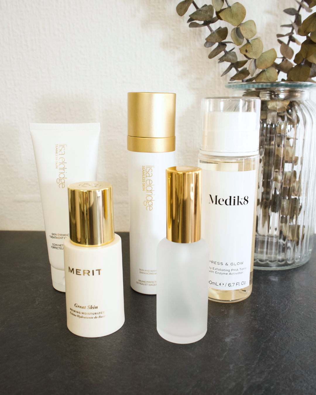 Collection of skincare products and a glass jar with dried eucalyptus leaves on a dark surface wall.