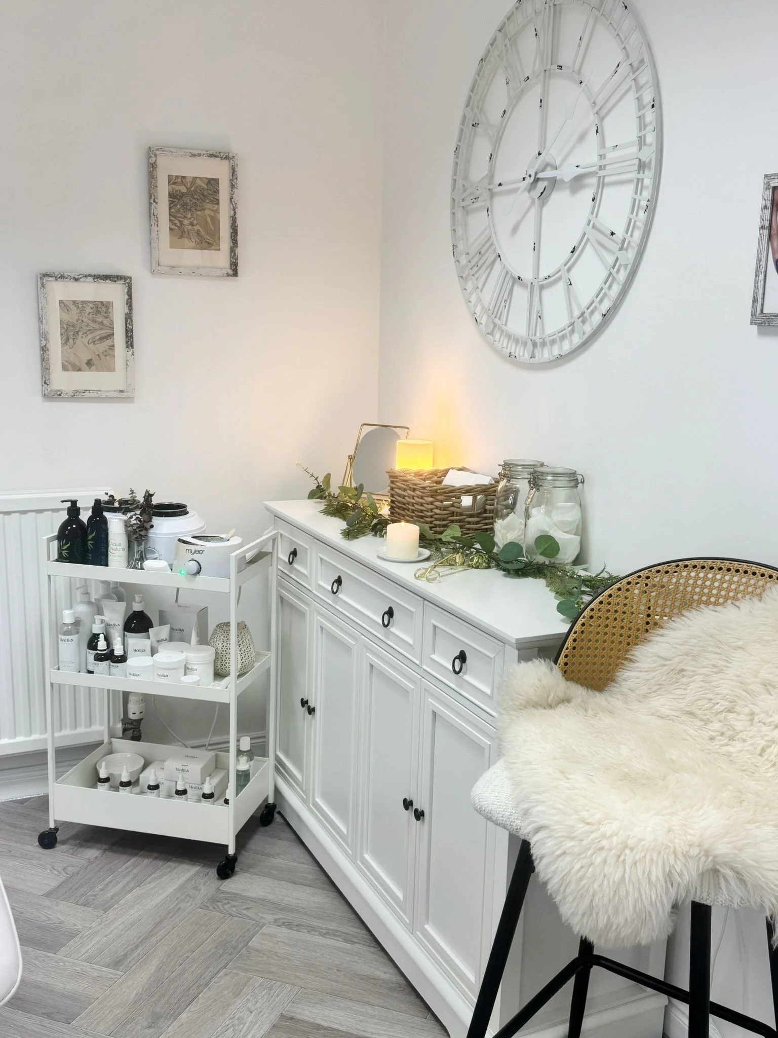 A cozy corner of a room with a white cabinet, a fluffy beige chair with black legs and a fur throw, a wicker basket, candles, glass jars, and skincare products on a trolley. A large white clock hangs on the wall and framed artwork decorates the space.