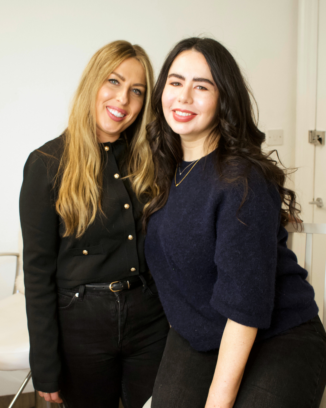 Two women smiling and posing together indoors, one with long blonde hair and black outfit, the other with long dark hair and navy sweater.