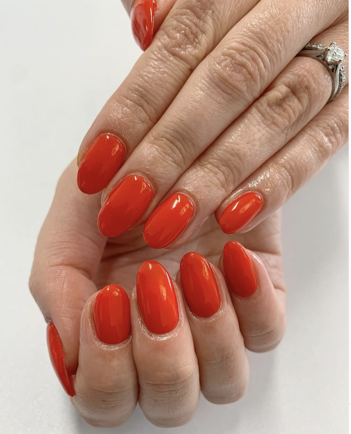 Close-up of a person's hand with bright orange-red painted nails, wearing a silver ring with a large clear gemstone.