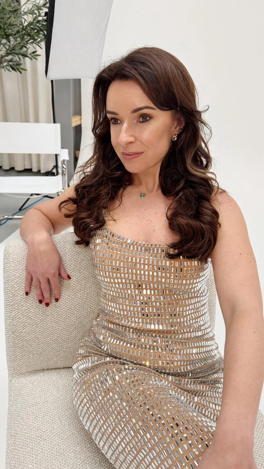 A woman with long, curly brown hair wearing a sleeveless, metallic gold dress with a grid pattern, sitting on a beige chair in a bright room with a white background and a potted plant.