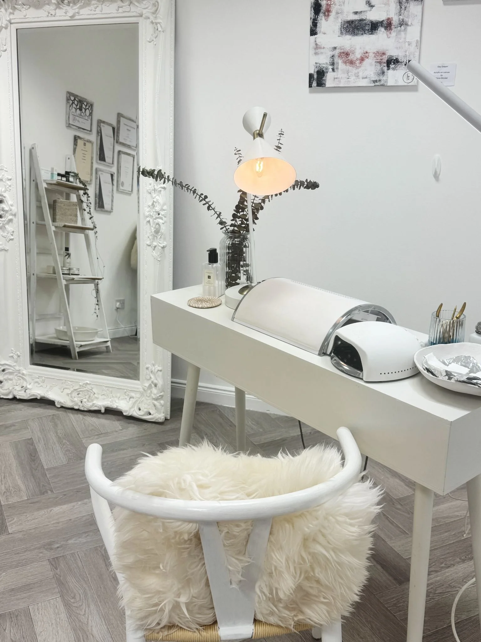 A cozy, decorated room with a large ornate white framed mirror, a ladder shelf with framed certificates or photos, a table with a decorative vase with eucalyptus and a lamp, a white nail polish bottle, a covered nail curing light, and a fluffy cream-colored chair with a rounded frame.