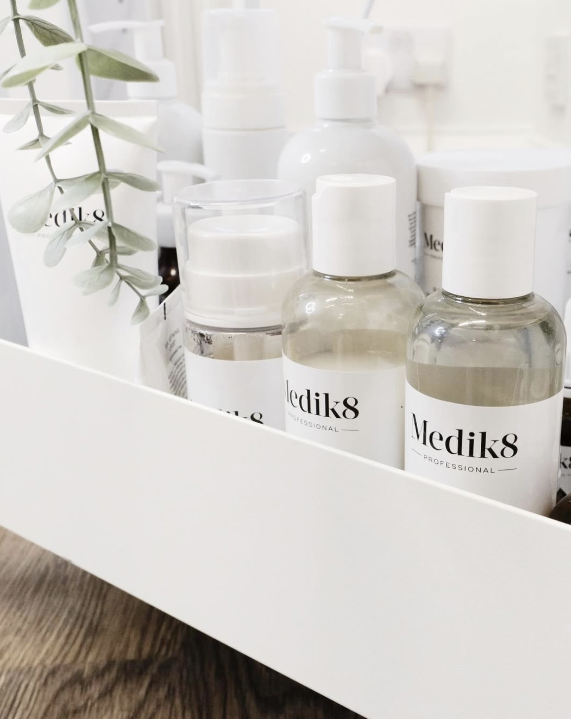 Collection of white bottles and containers with black and white labels, some with clear liquid and a sprig of green leaves in a white storage box.
