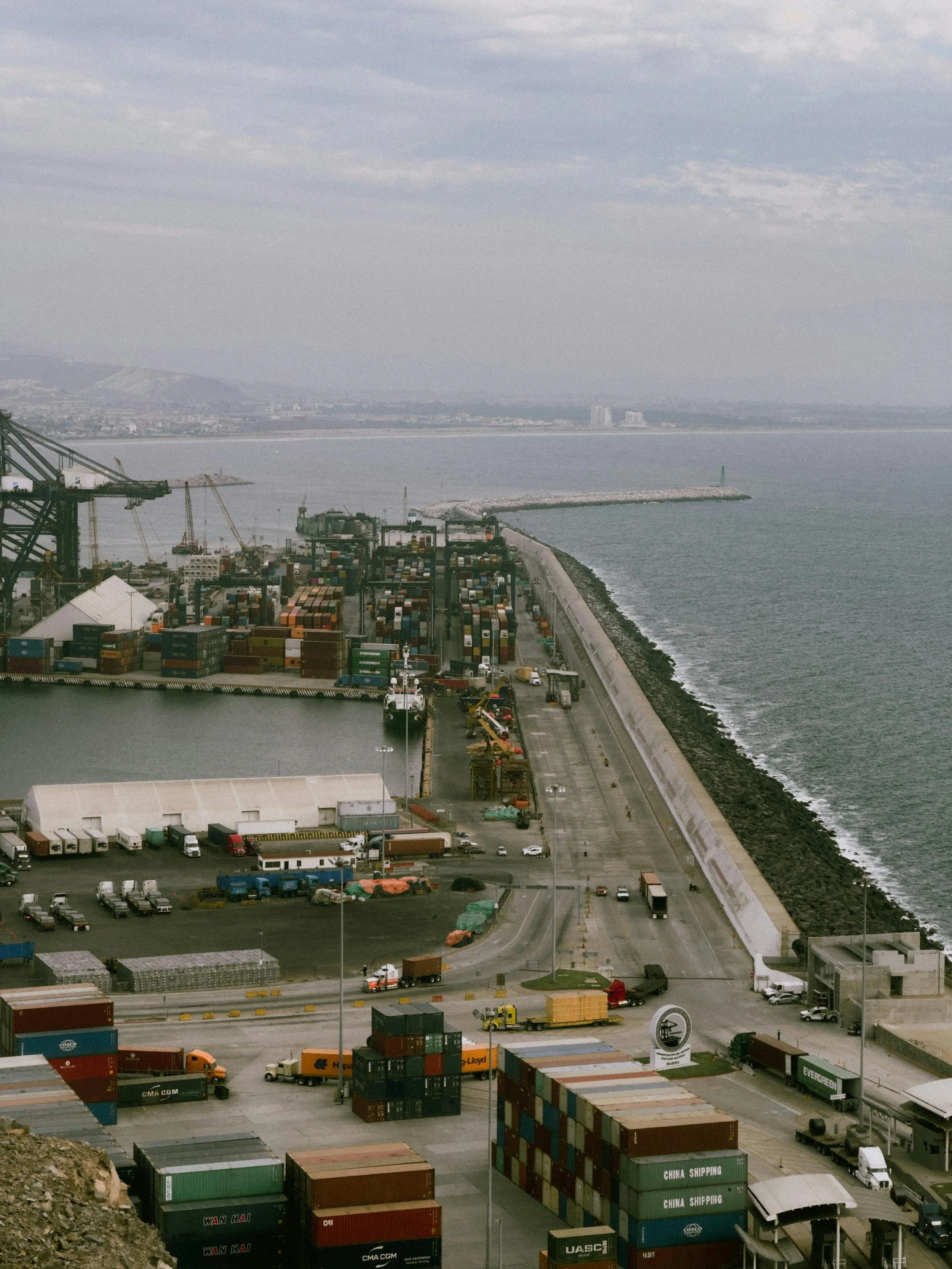 View of a busy port with numerous stacked shipping containers, trucks, and cranes, overlooking the ocean with a breakwater and distant landmass under cloudy skies.