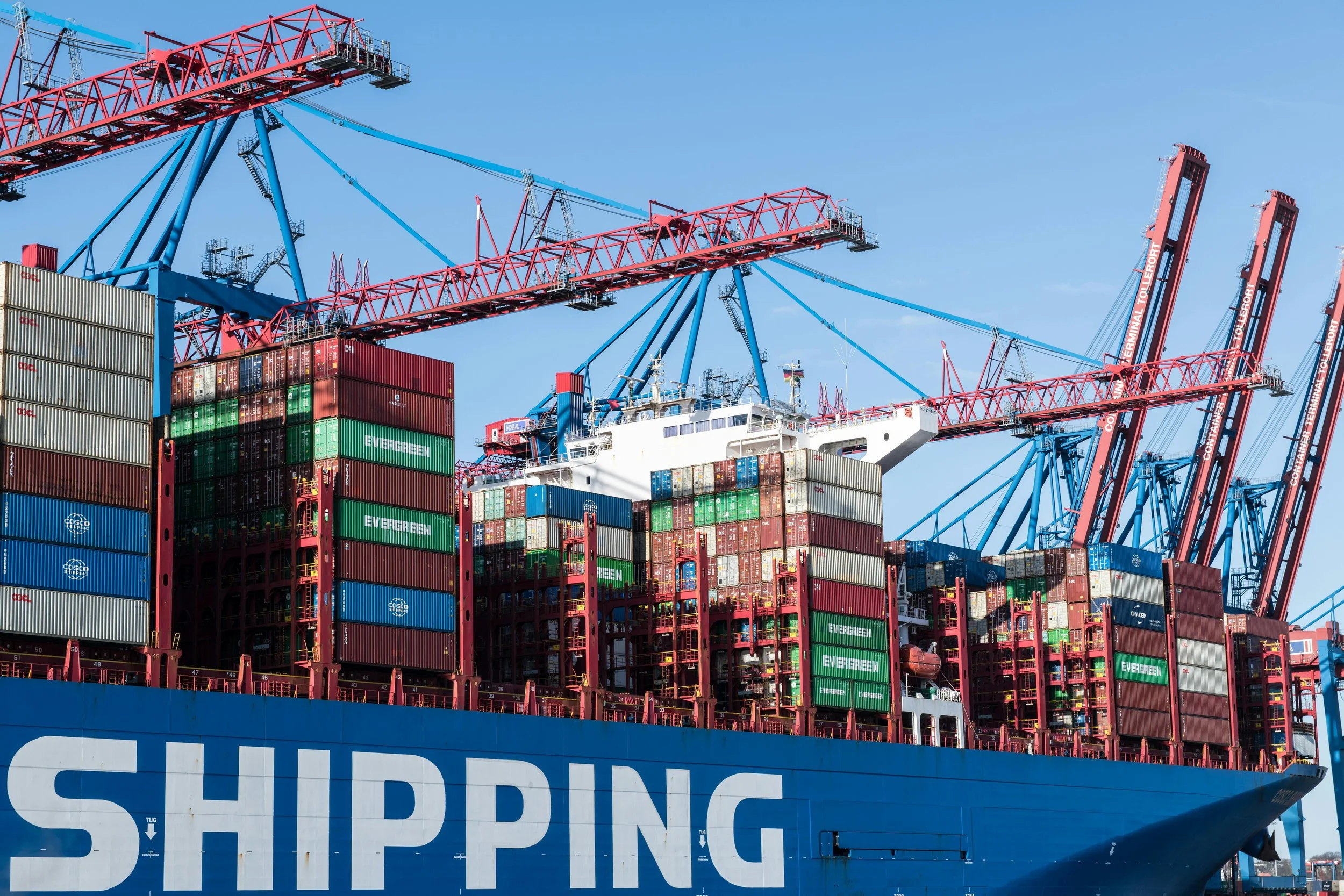 Container ship with stacks of multicolored shipping containers and large red and blue cranes against a clear blue sky.