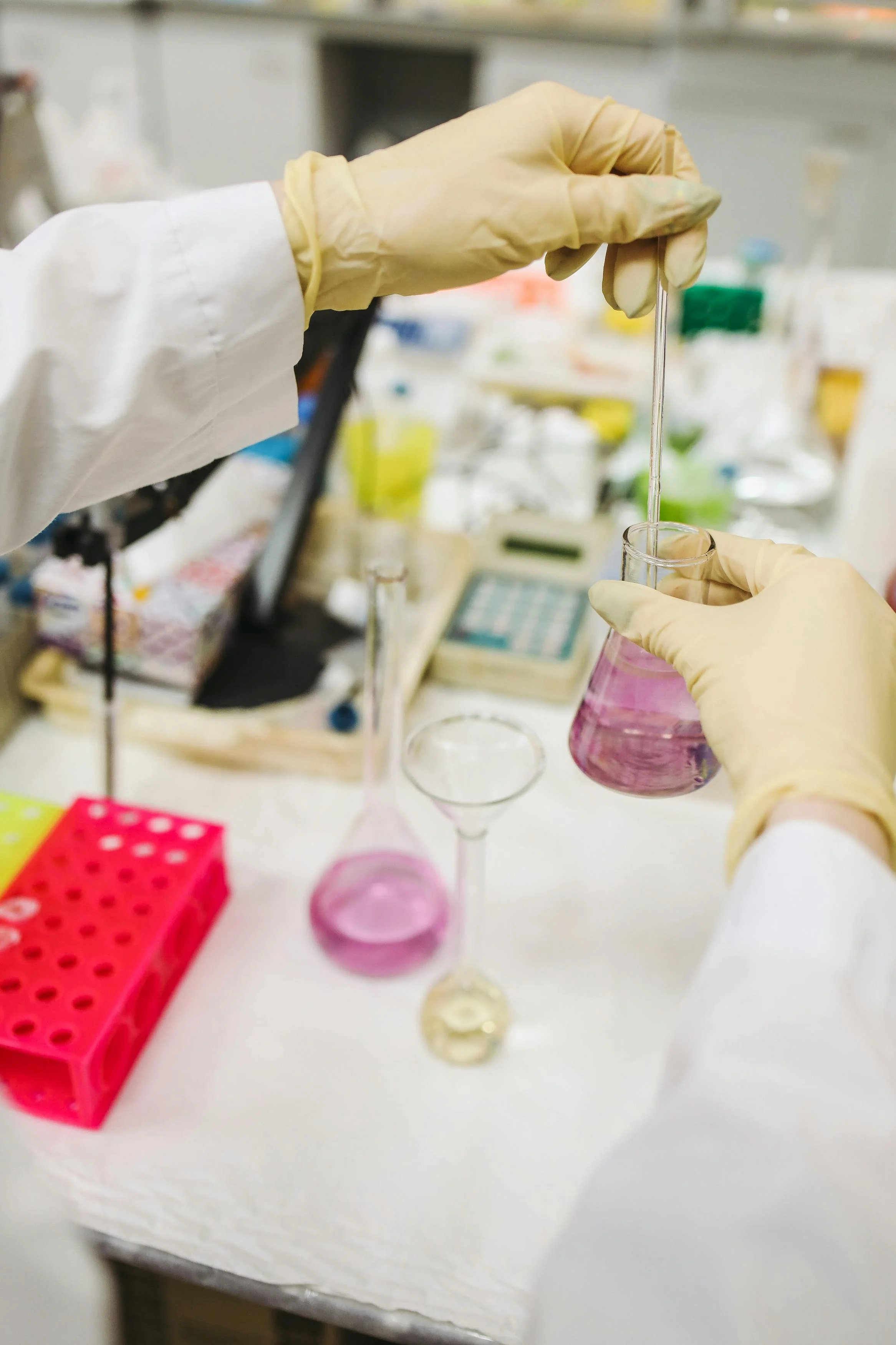 A scientist wearing gloves and a lab coat is using a pipette to transfer liquid into a graduated flask in a laboratory setting.