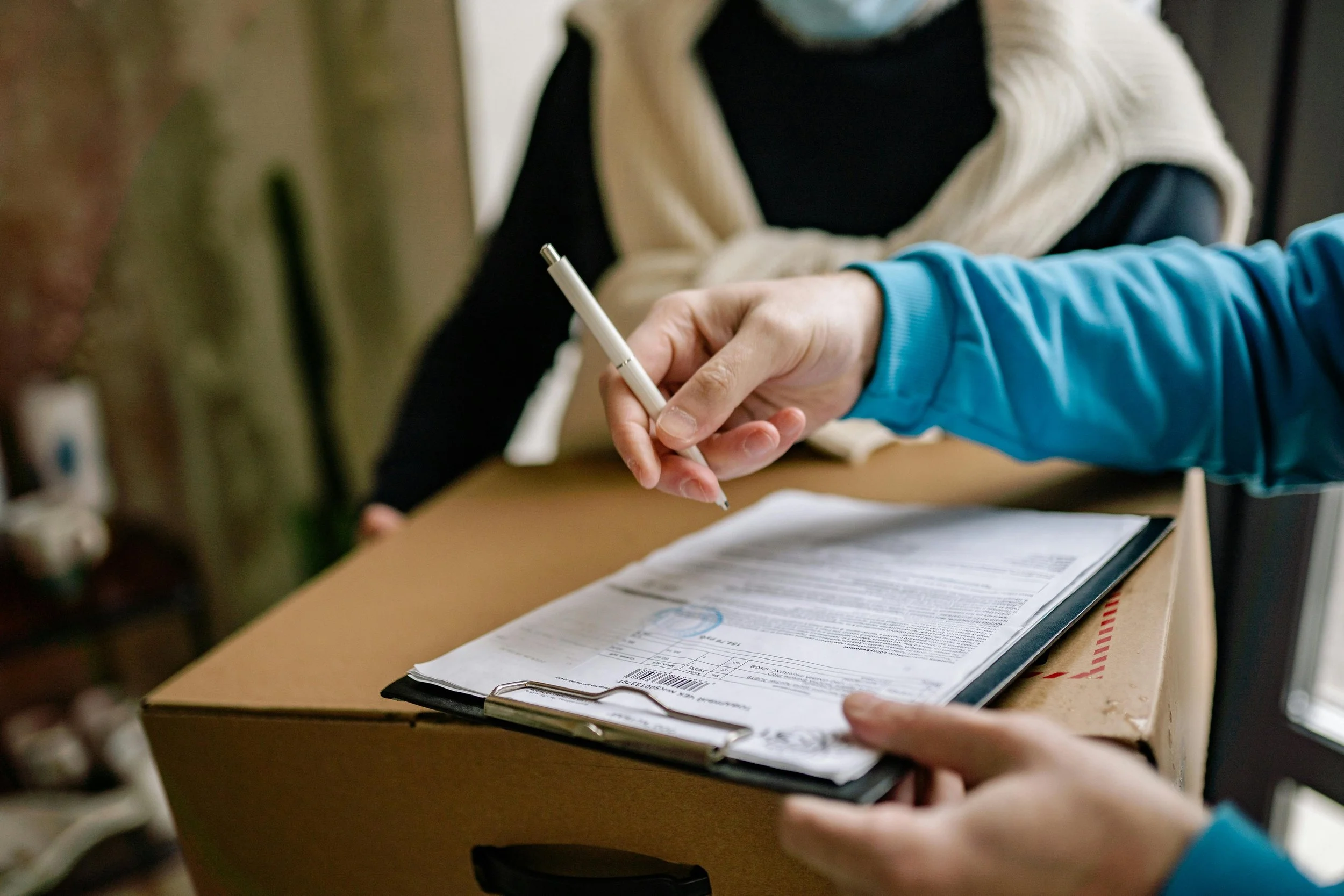 Person holding a clipboard with papers, sitting at a table, writing with a pen.