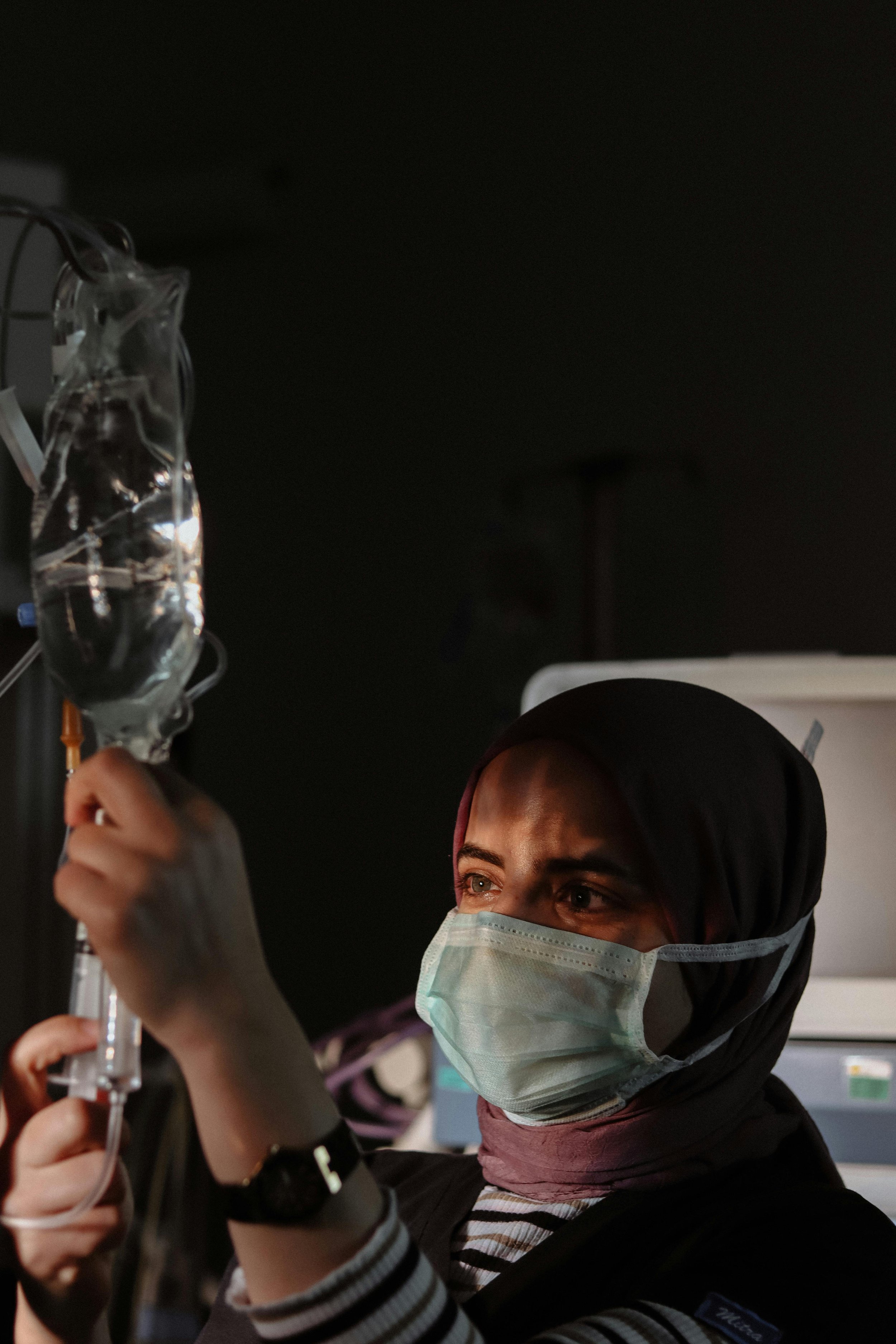 A person wearing a face mask is holding a syringe with a liquid, likely preparing a vaccination or injection. The background is dark and the person's eyes are visible above the mask.