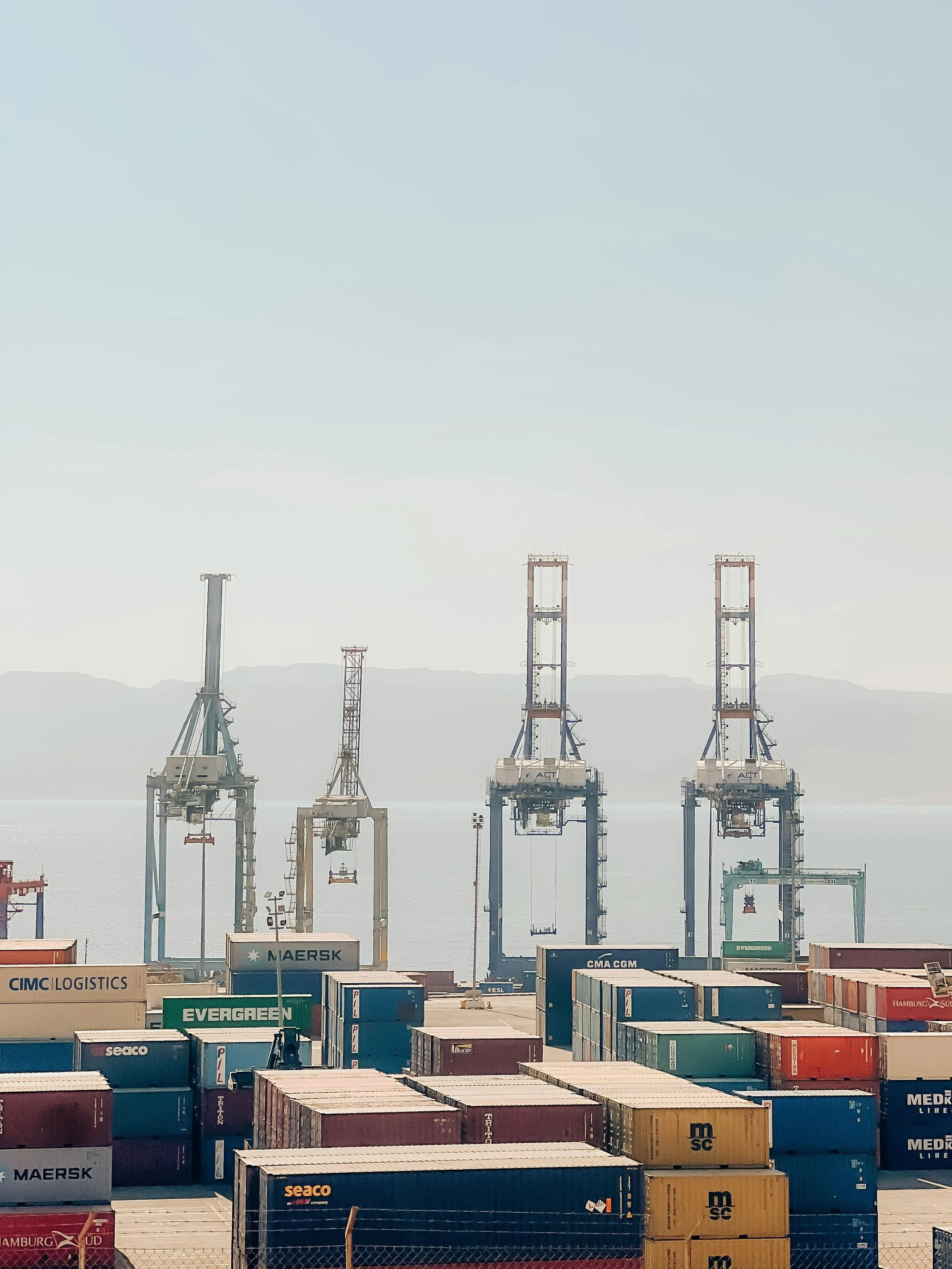 Cargo containers at a port with crane structures in the background and mountains visible in the distance.