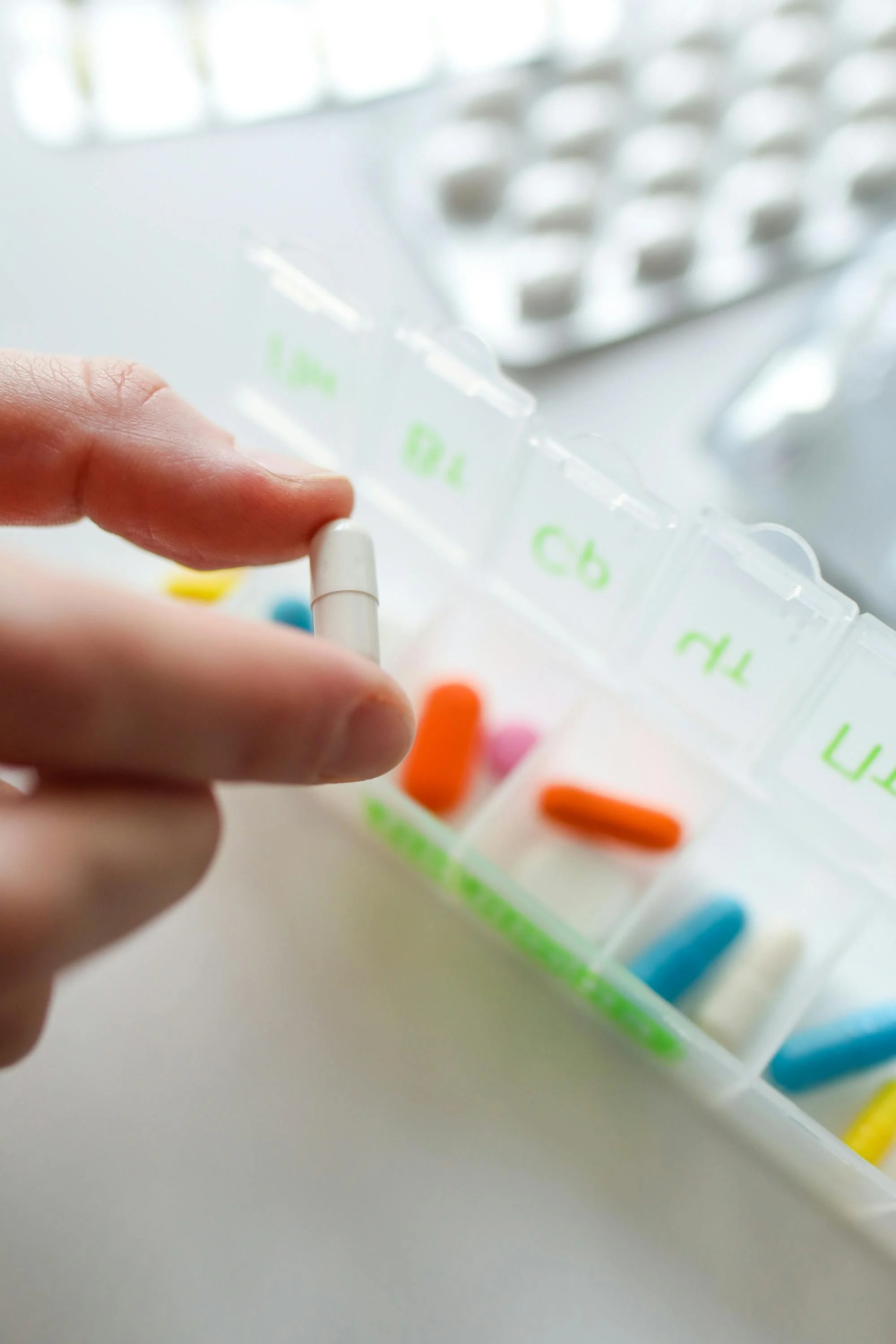 Close-up of a person's hand holding a white pill against a background of colorful capsules and a pill organizer.
