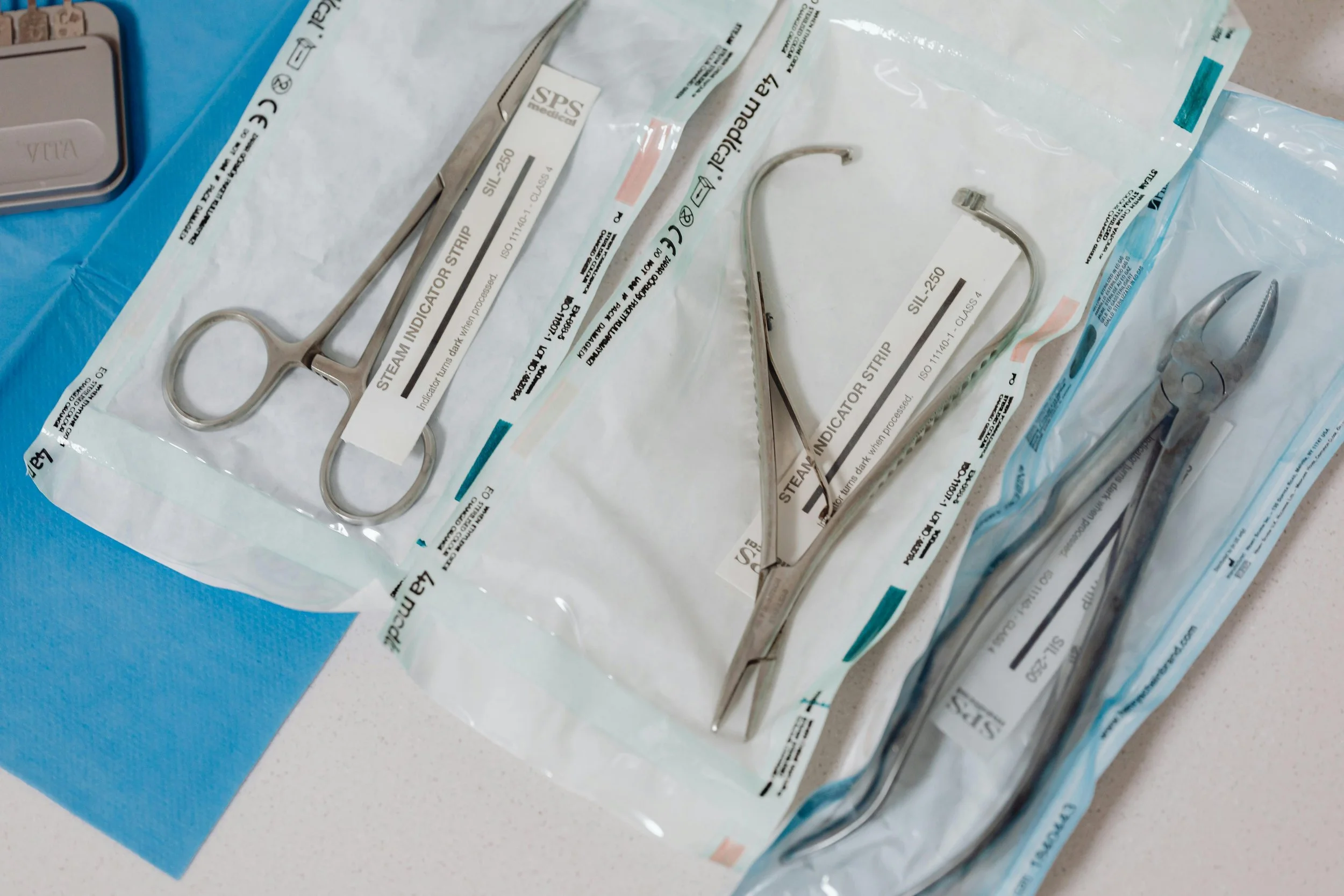 Sterile medical tools, including a pair of forceps and a small curved surgical instrument, are laid out on sterile packaging on a medical table.