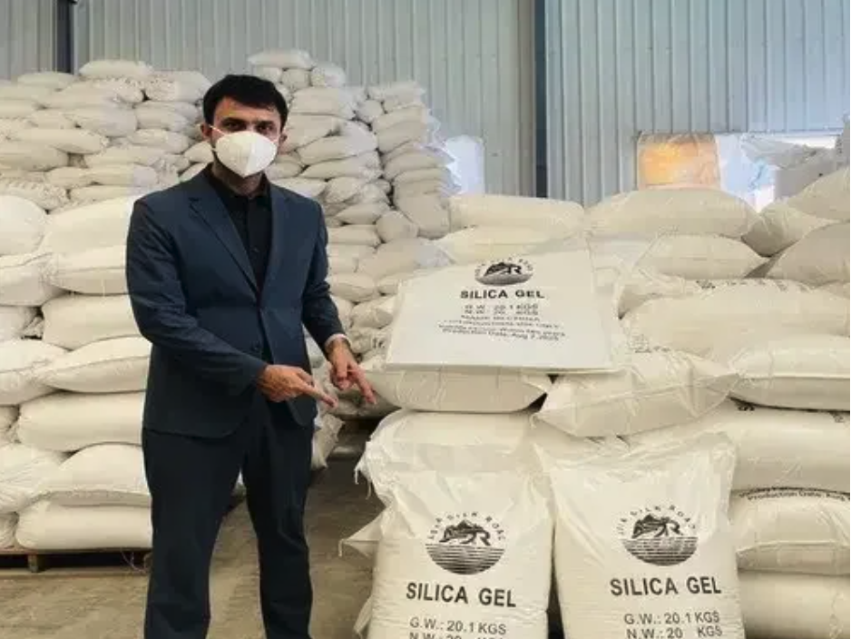 Man in business attire and face mask standing next to large bags of silica gel inside a warehouse.