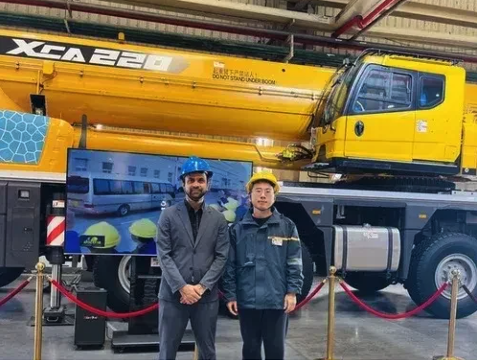 Two men in safety helmets standing in front of a large yellow crane inside a factory or warehouse.