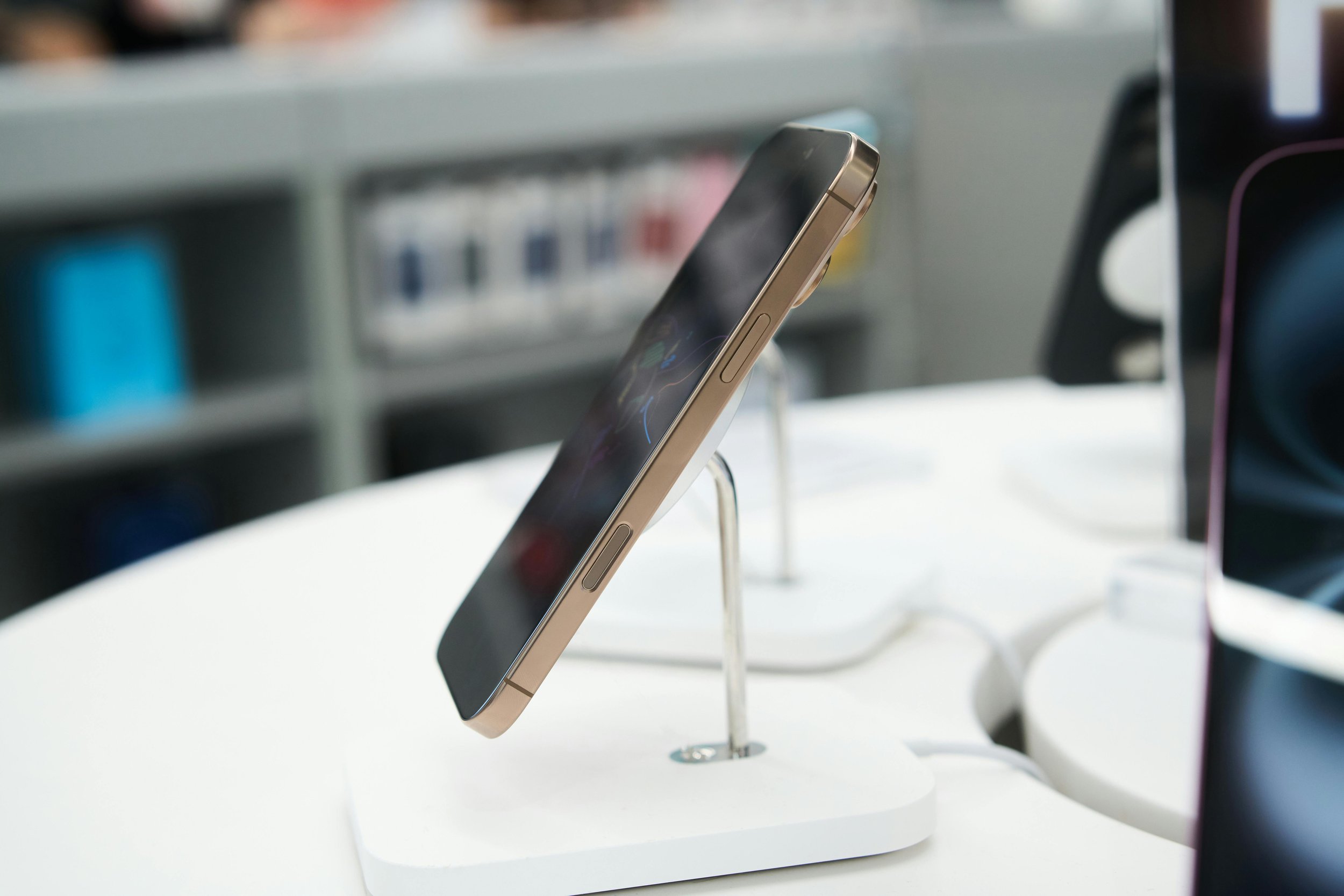A smartphone displayed on a white stand in a store, with shelves of other electronic products blurred in the background.