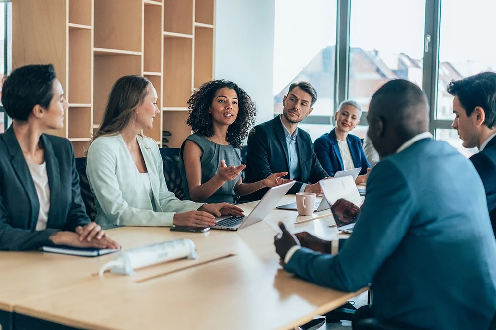 A diverse group of professionals in a business meeting or discussion around a conference table with laptops and notebooks in a modern office.