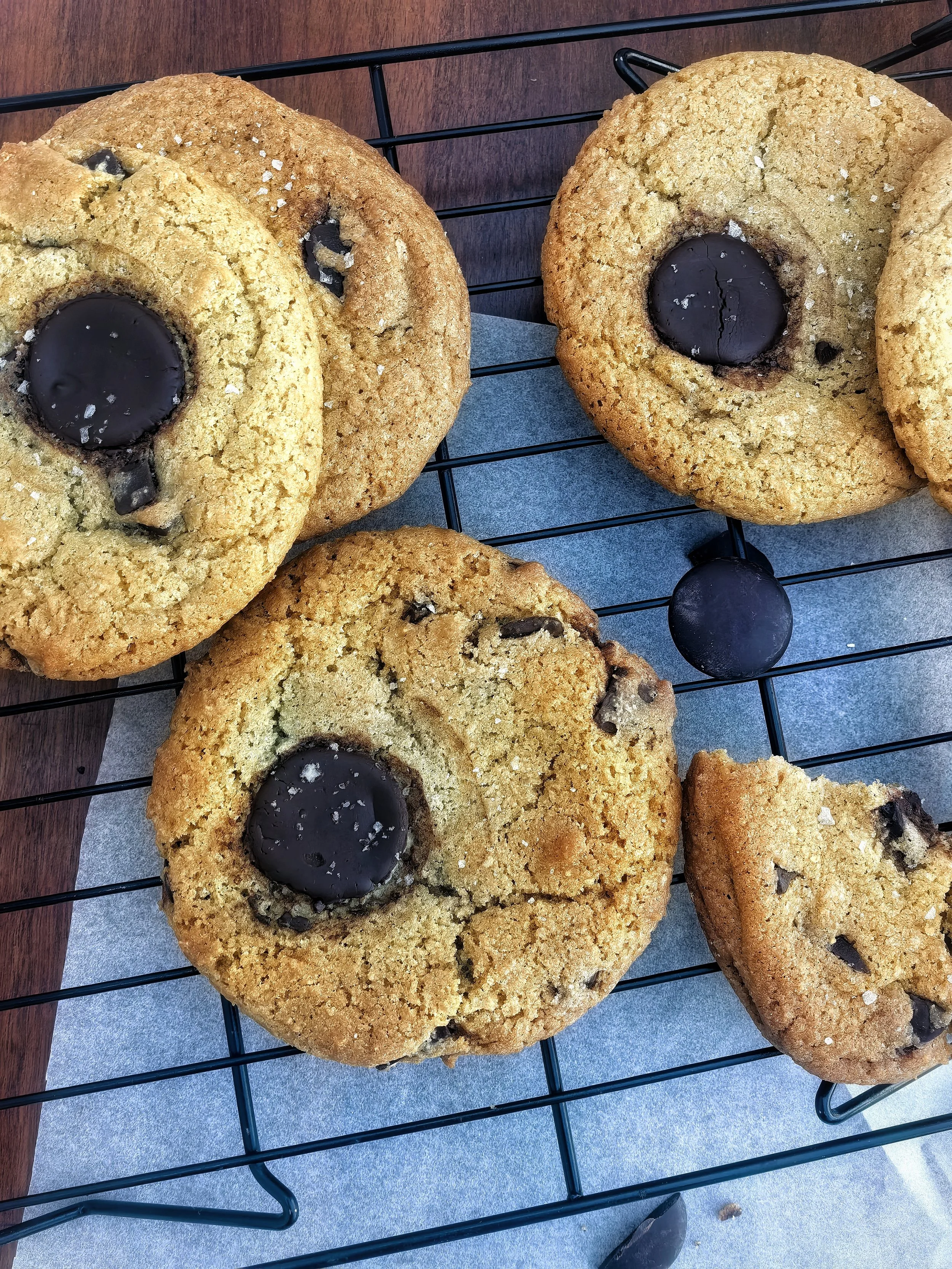 Close-up of three large chocolate chip cookies with salt sprinkled on top, resting on a cooling rack over blue parchment paper.