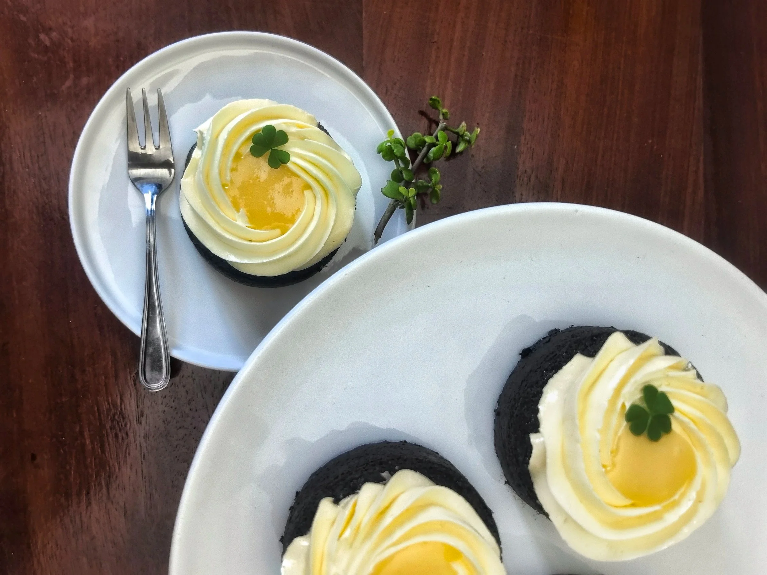 Close-up of  chocolate mini-cake with white filling and lime-colored swirl. A fork rests on the plate, and there is a sprig of greenery on the side. The background features a wooden surface and a white bowl.