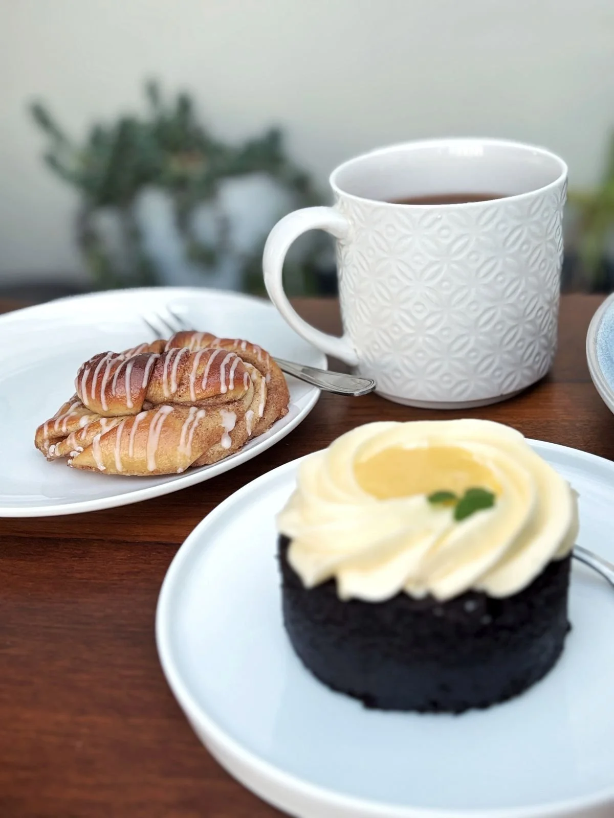 A plate with a chocolate mini-cake topped with white frosting and a green decoration, a plate with cinnamon rolls drizzled with white icing, a white mug with hot tea.