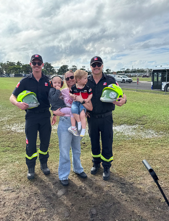 Two firefighters in uniform standing outdoors with a woman and two young children, one of the children wearing headphones, at a public event on a cloudy day.