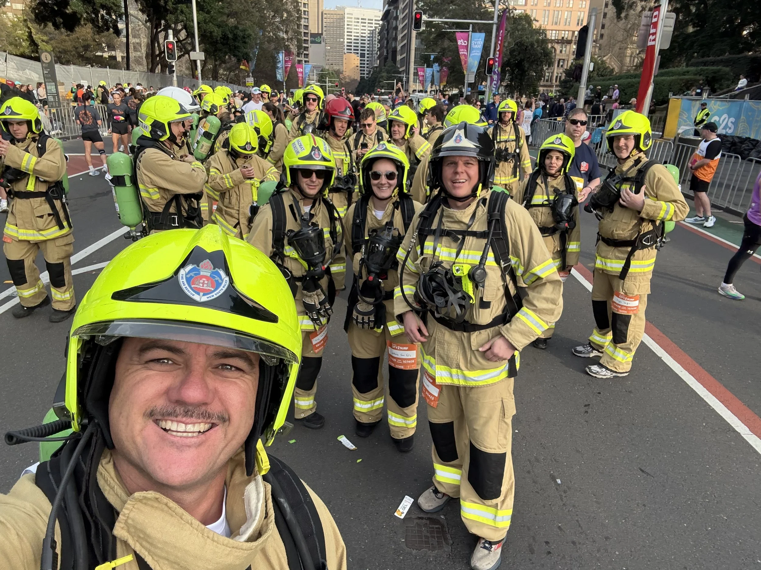 A man wearing a fire helmet taking a selfie in front of a group of firefighters dressed in tan uniforms with yellow helmets, at an outdoor event in an urban area with tall buildings and street lights.