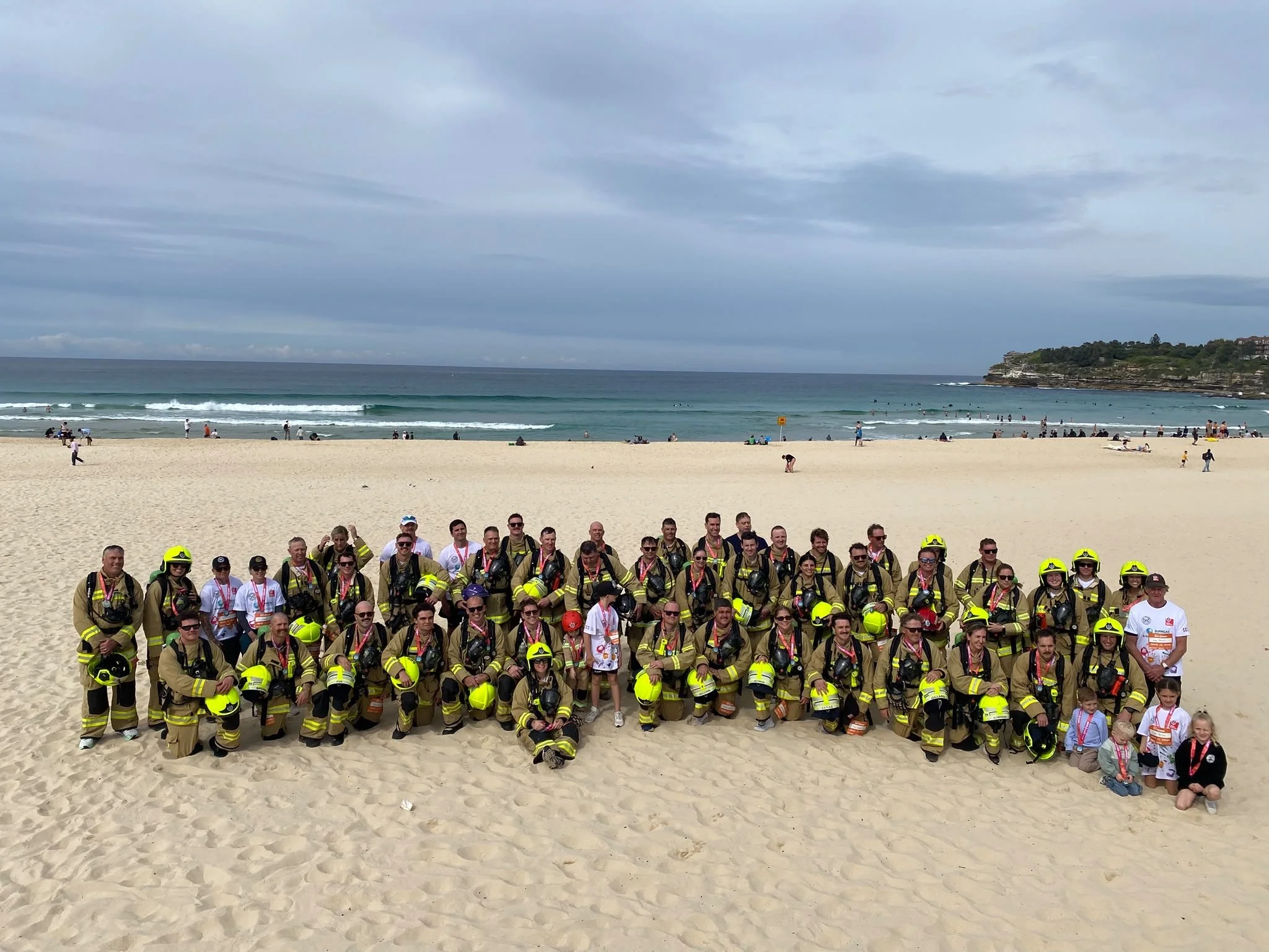 A large group of people, including firefighters in yellow and black uniforms and helmets, posing for a photo on a sandy beach. The background features an ocean with waves and a cloudy sky.