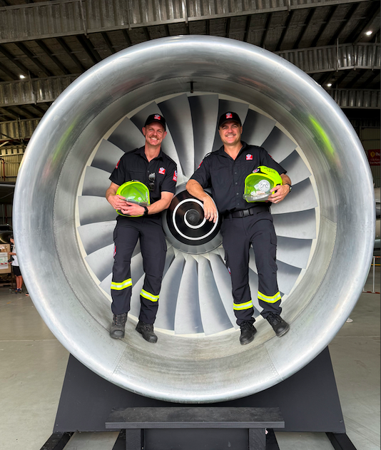 Two firefighters in uniform standing inside a jet engine, holding green helmets, smiling for the photo.