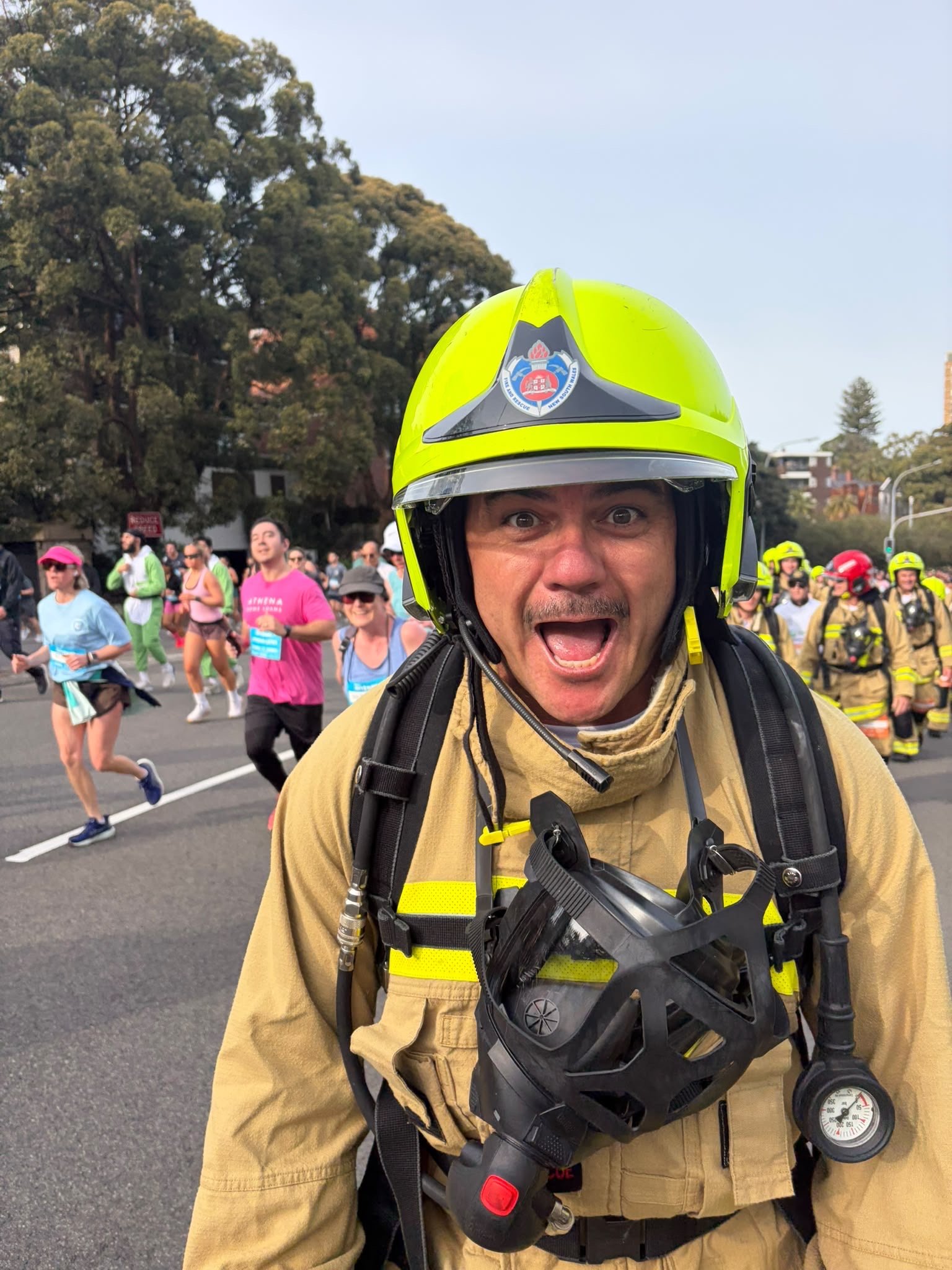 A firefighter in uniform and helmet on a street during a marathon, with runners participating in the race visible in the background.