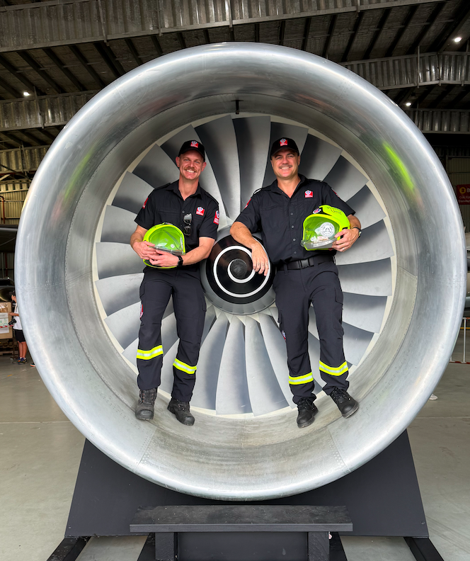 Two firefighters in uniform standing inside a large aircraft jet engine, holding green helmets and smiling.