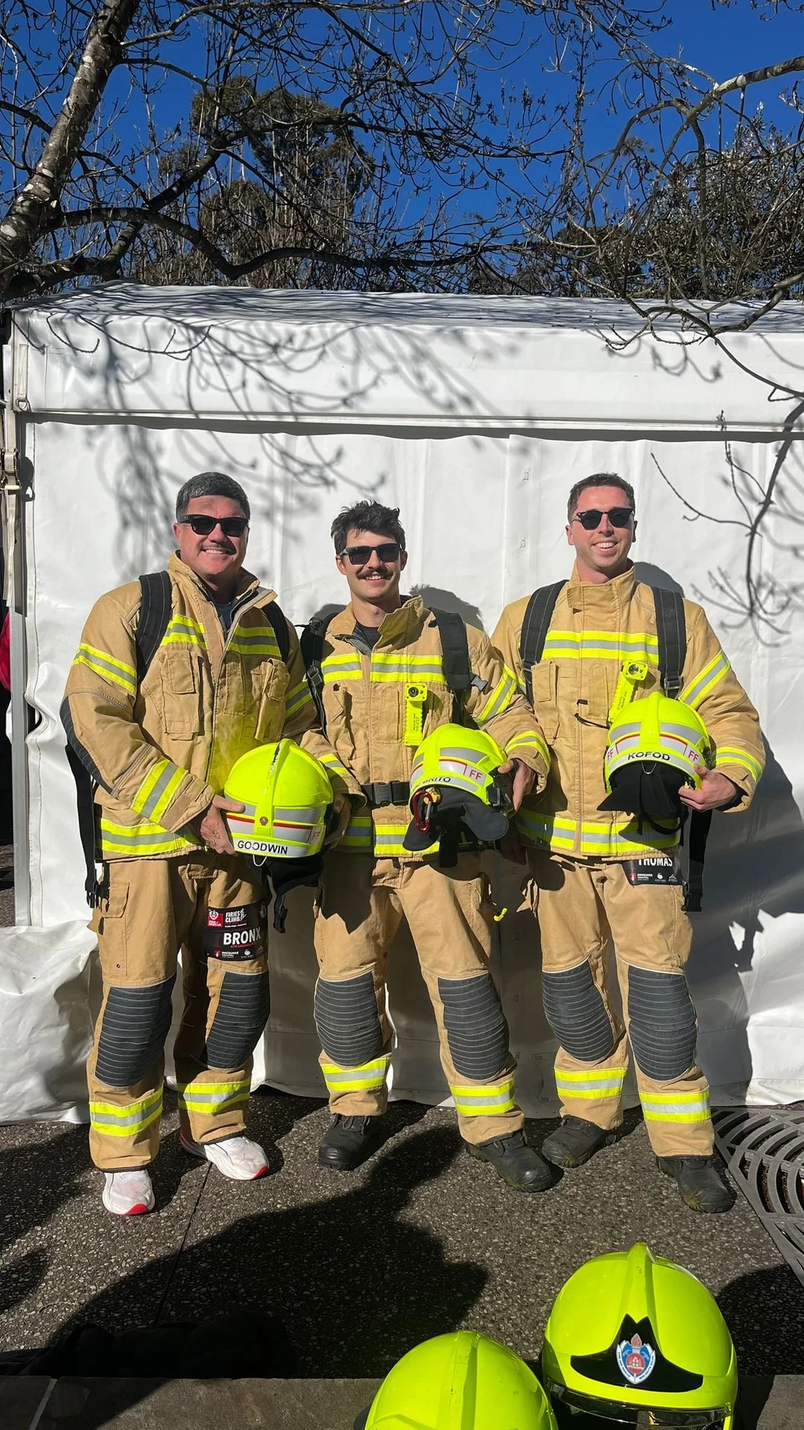 Three firefighters in tan firefighting gear standing outdoors in front of a white tent, holding yellow helmets, with leafless trees and a blue sky overhead.