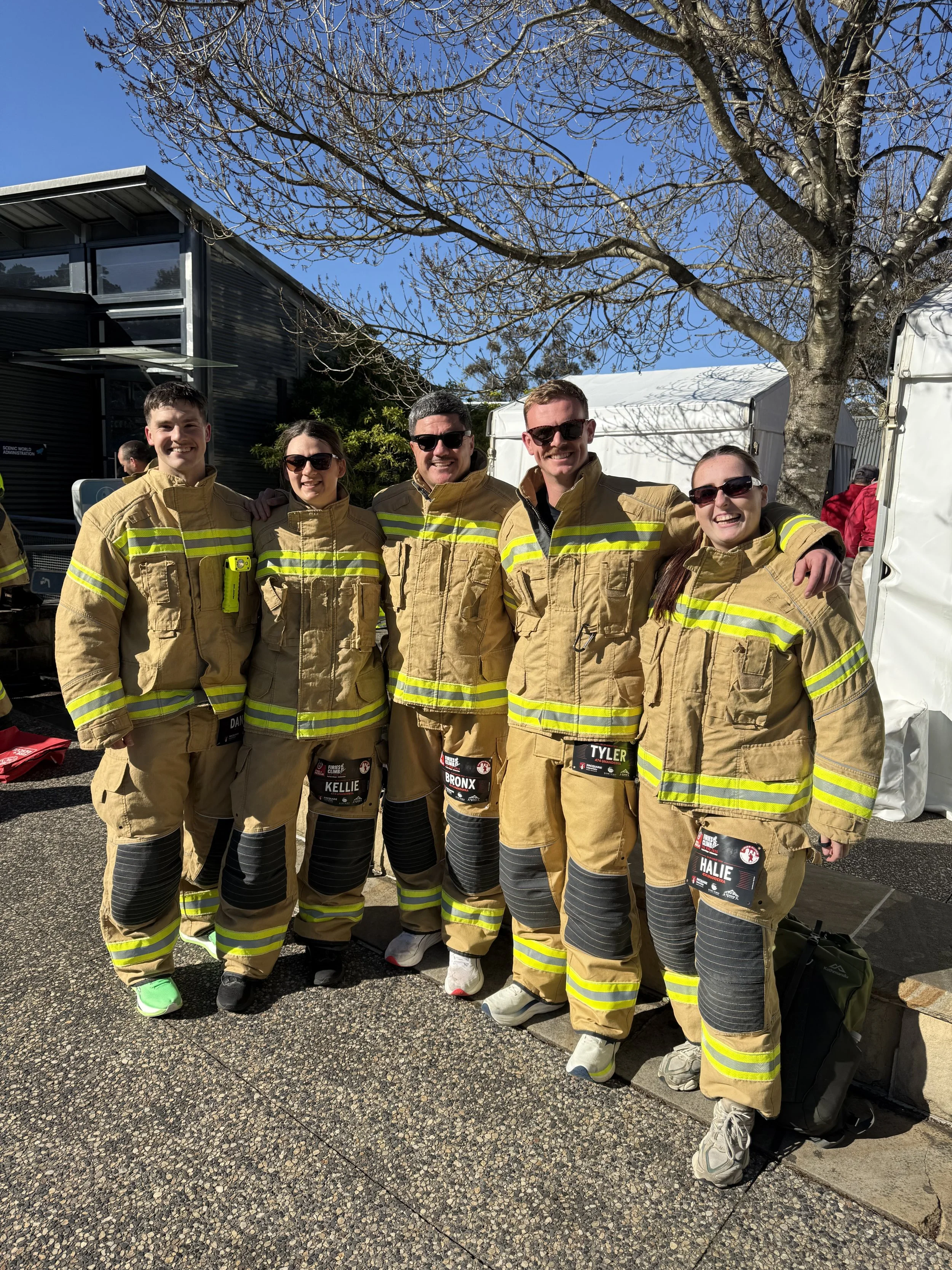 Group of five firefighters in tan uniform with yellow reflective stripes standing outdoors under a leafless tree, smiling at the camera.