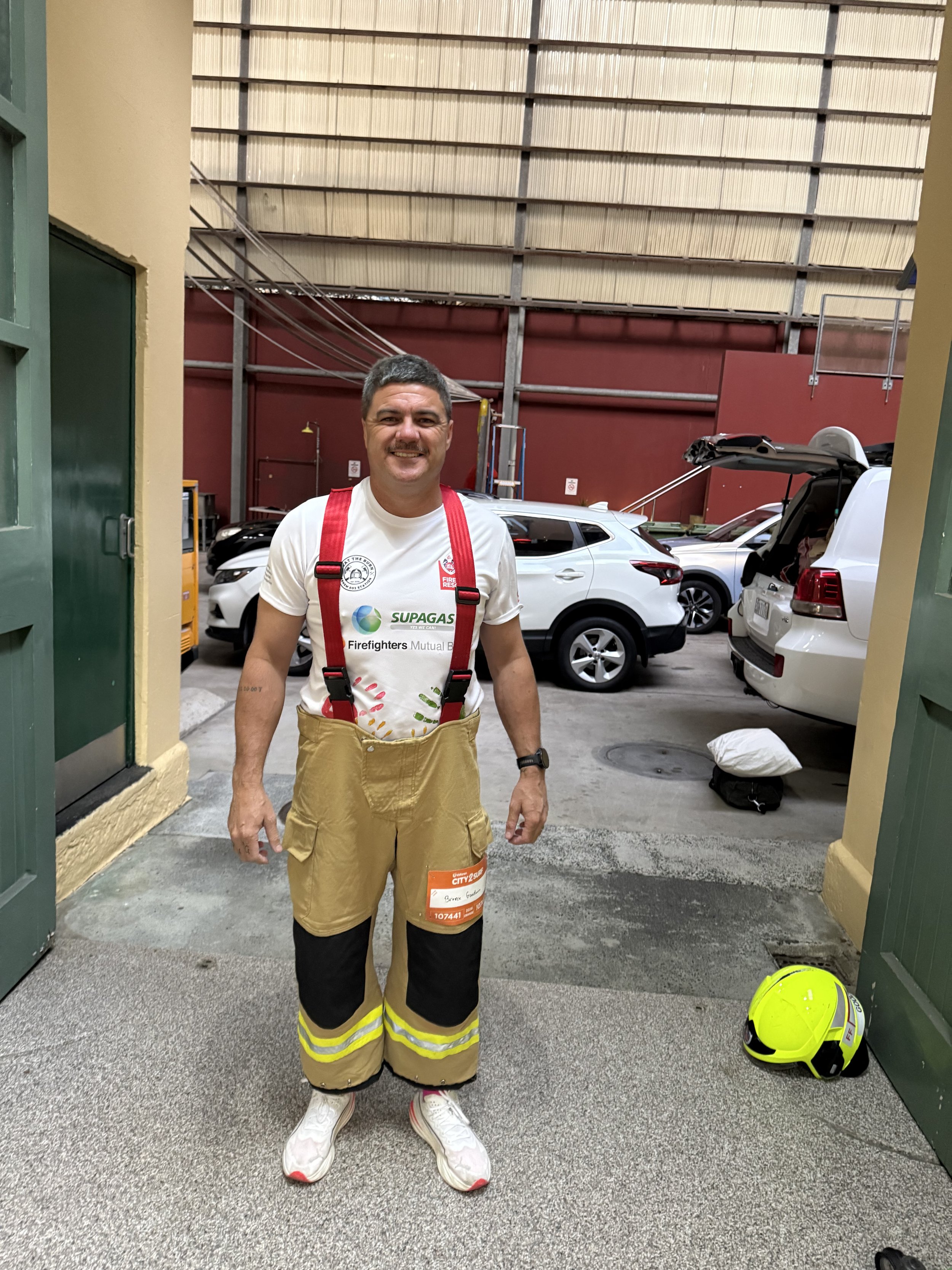 A firefighter stands outdoors in front of parked cars, smiling at the camera. He is wearing firefighter gear, including fire-resistant pants with a reflective stripe, sneakers, and a white t-shirt with various logos. A yellow firefighter helmet is on the ground to his right.