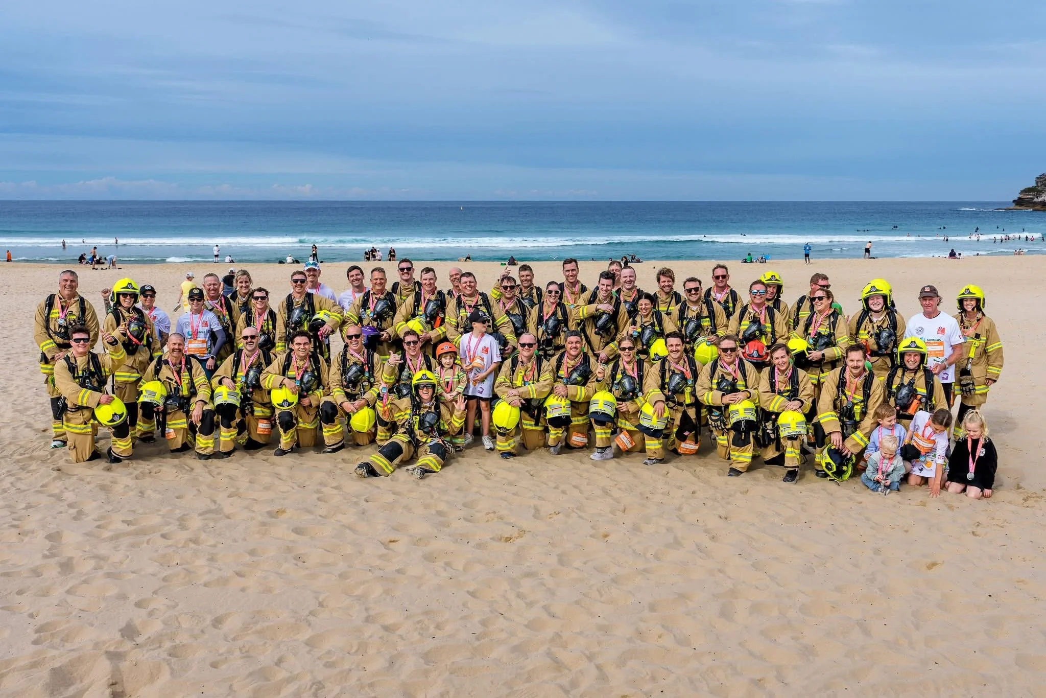 A large group of firefighters in uniform, some wearing helmets, standing together on a sandy beach with the ocean and a cloudy sky in the background.