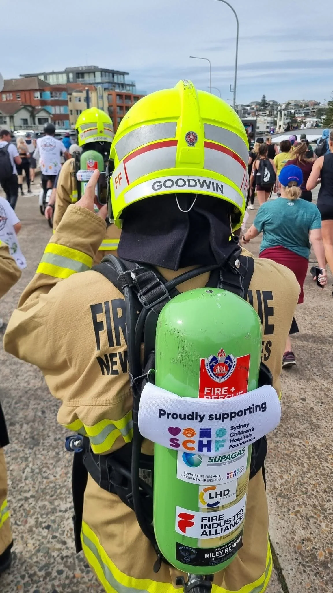 A firefighter wearing a yellow helmet with the name 'Goodwin' and a beige uniform with yellow reflective stripes, carrying a green oxygen tank supported by a harness, with multiple rescue and firefighting organization logos and supporting messages, amidst a crowd at an outdoor event.