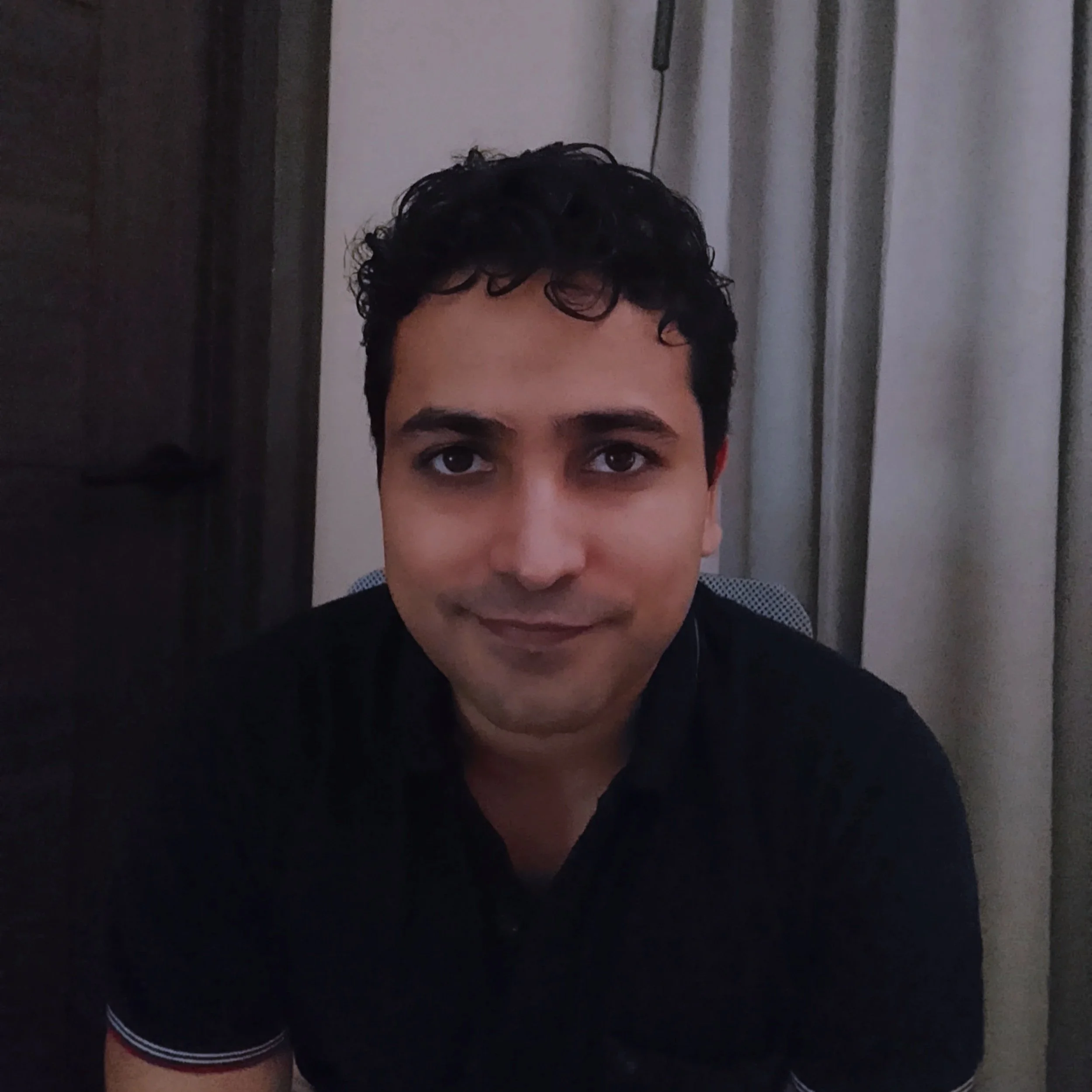 Partial view of a person with dark, curly hair and eyebrows, wearing a dark shirt with a small white pattern.