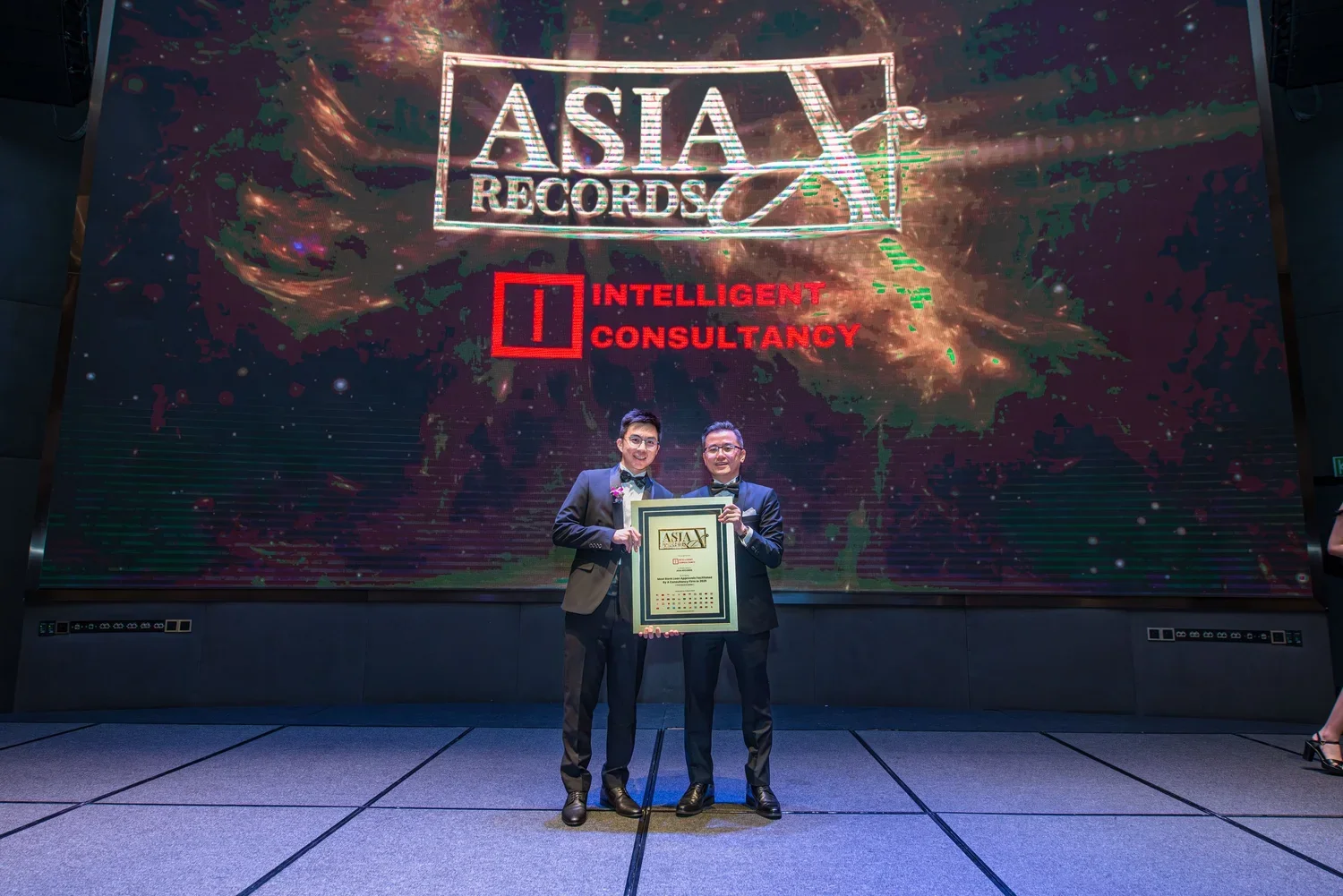 Two men in tuxedos standing on a stage, holding a framed award, in front of a large digital screen displaying 'Asia Records' and 'Intelligent Consultancy' at an awards ceremony
