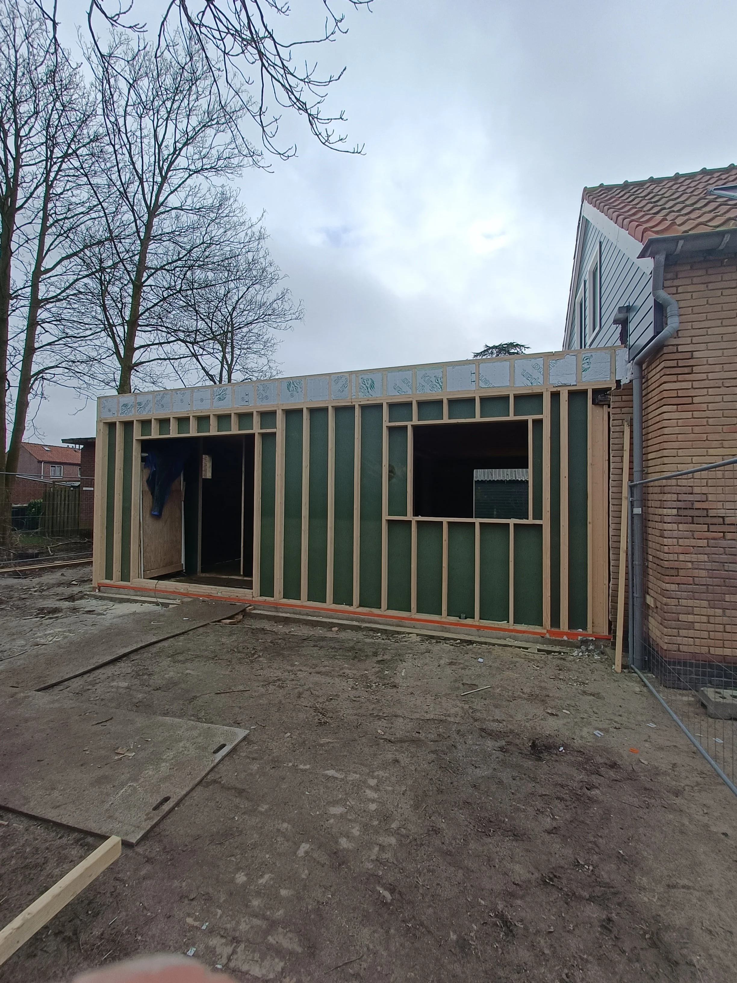 A house under construction with an unfinished wooden structure, showing vertical framing studs, against a background of leafless trees and an overcast sky.