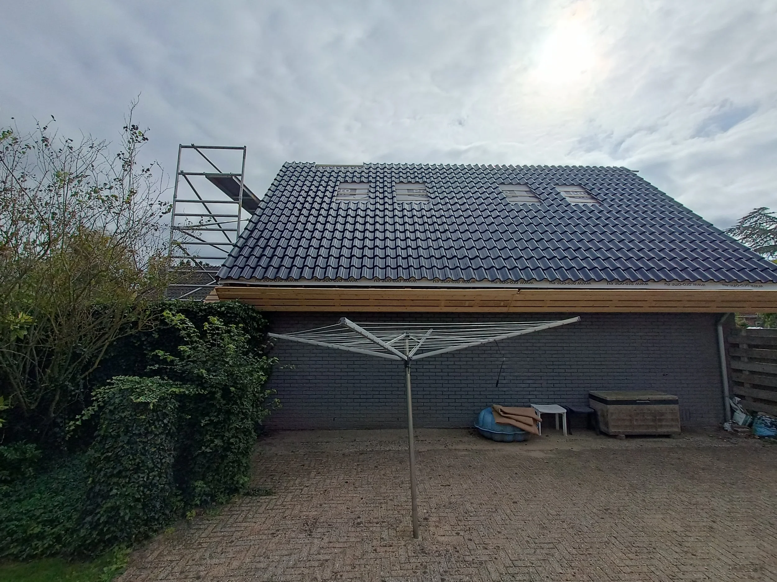 A house with a newly installed tiled roof, scaffolding on the left side, and a gray sky overhead. There is a brick wall and outdoor items including a blue boat and a folding chair in the yard.