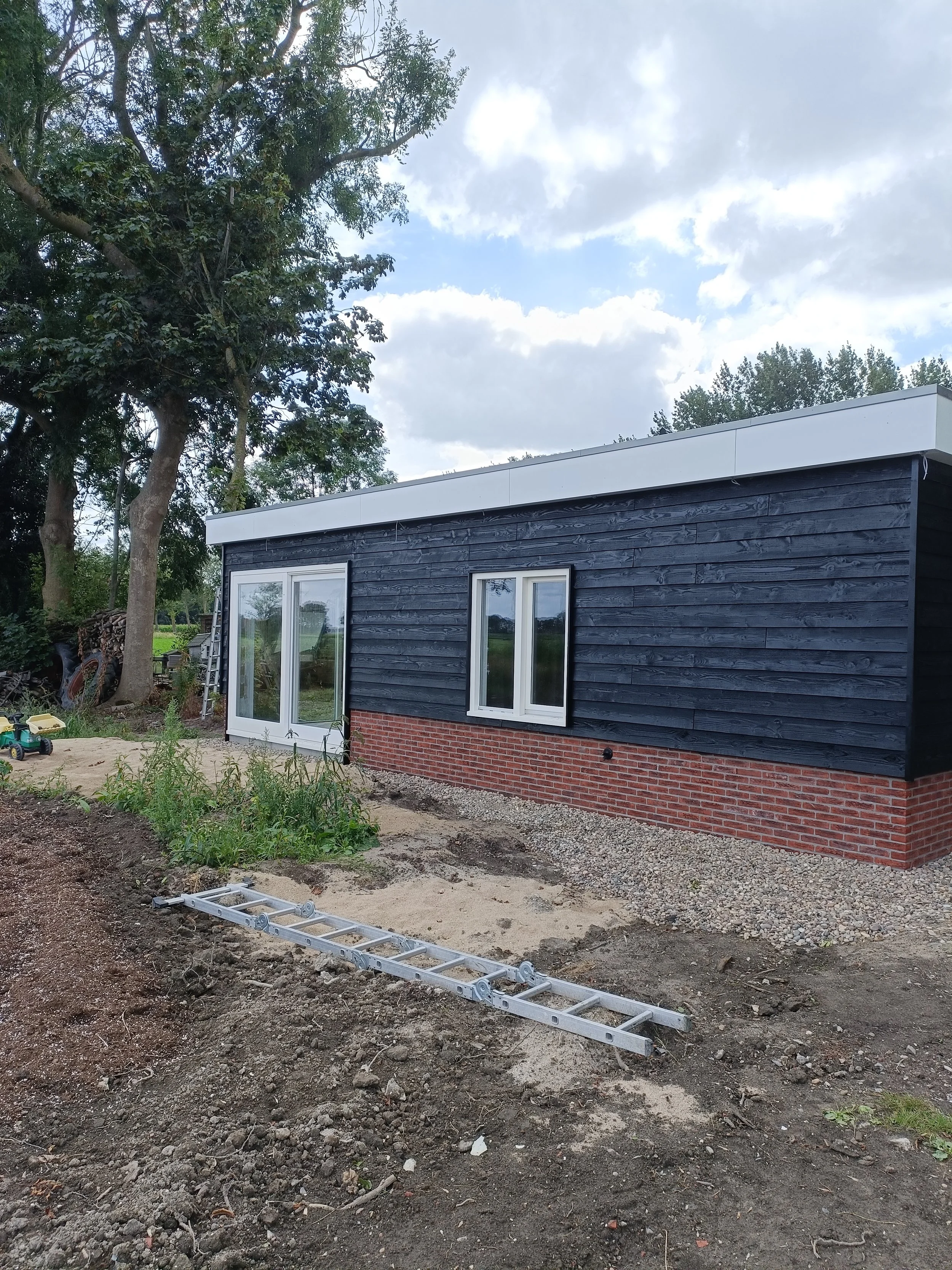 A partially constructed building with black wooden siding and white-framed windows, set on a brick foundation. The area around the building has exposed dirt and scattered soil, with some gardening tools and a ladder lying on the ground. Large trees and bushes are visible in the background, and the sky above is partly cloudy.