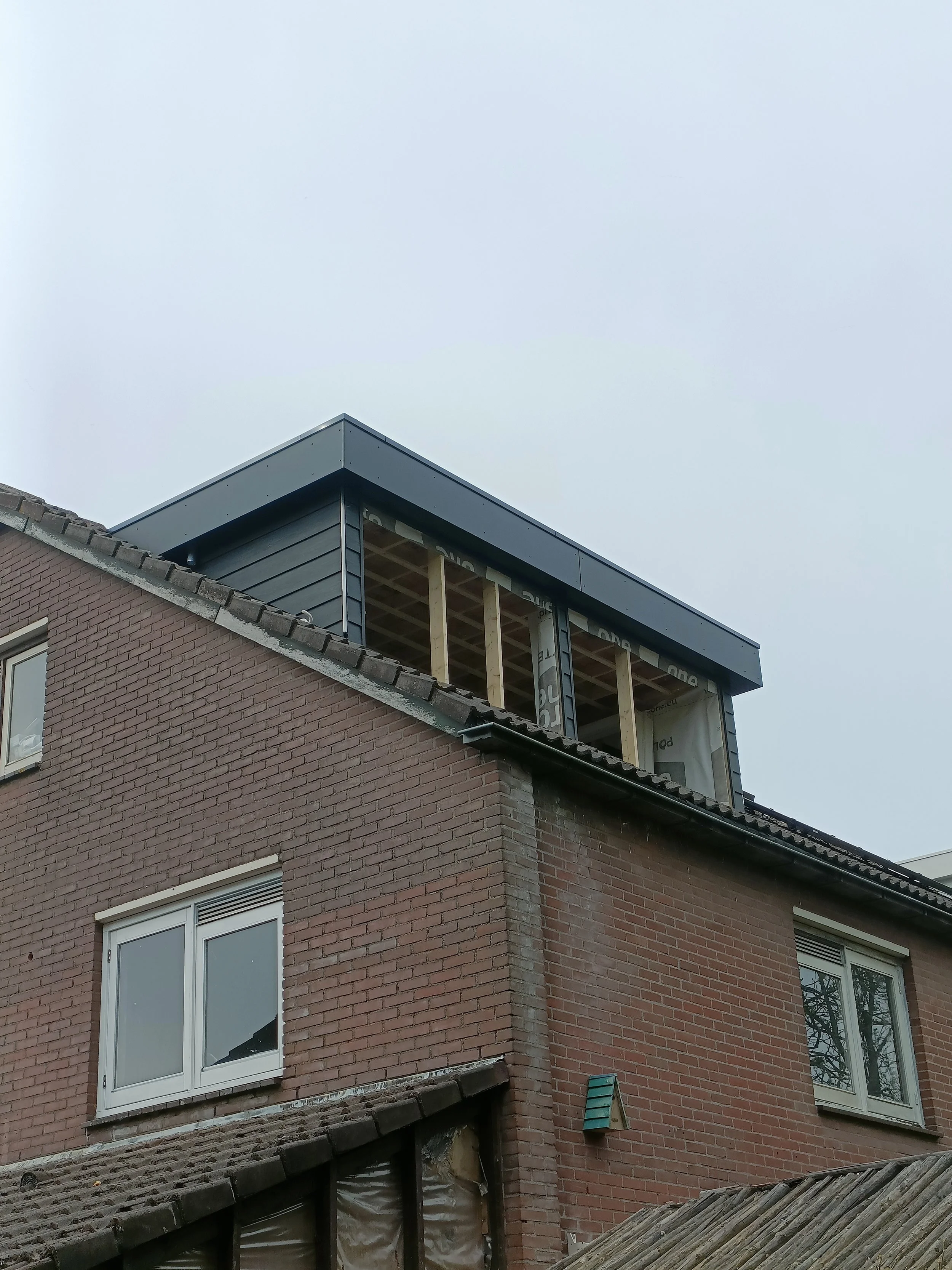 A brick residential building under renovation with an unfinished upper floor, exposed wooden beams, and an overcast sky.