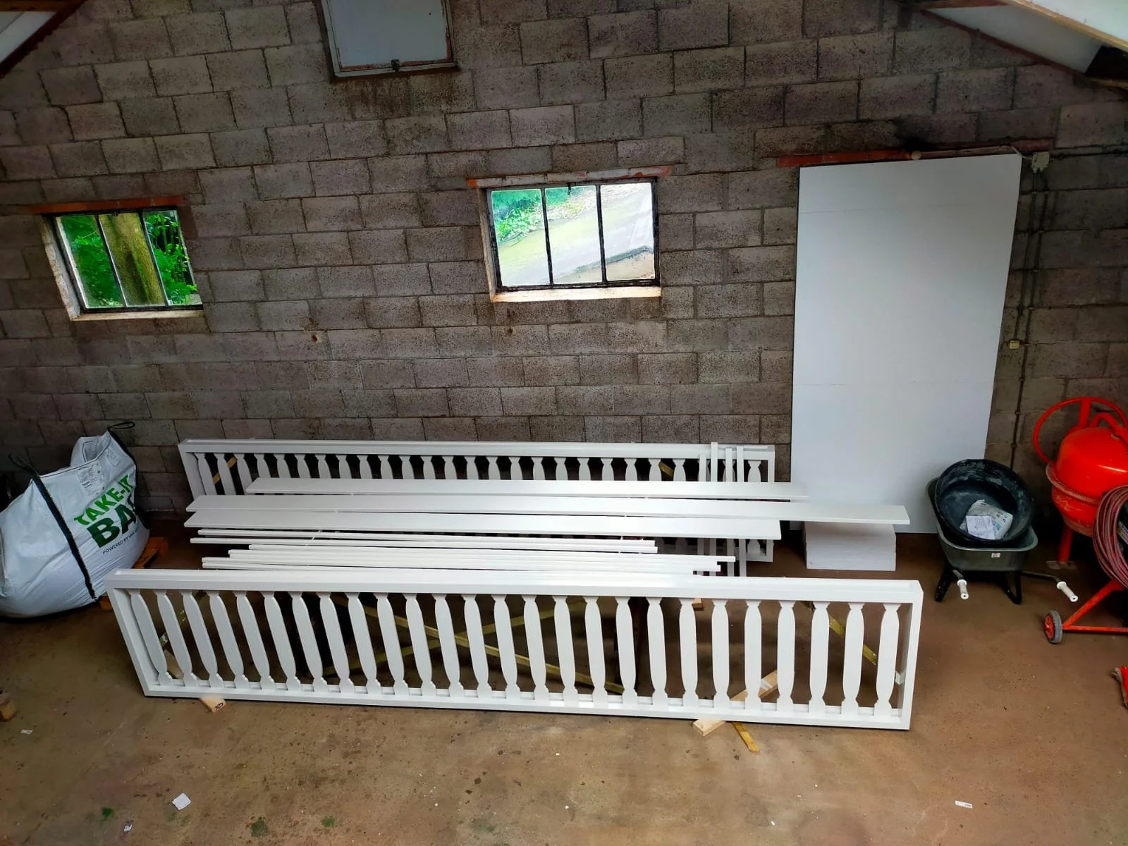 Storage room with unfinished cinder block walls, two small windows, white railing pieces, a black wheelbarrow with papers, a plastic container, a gas can, coiled garden hoses, and a large white board leaning against the wall.