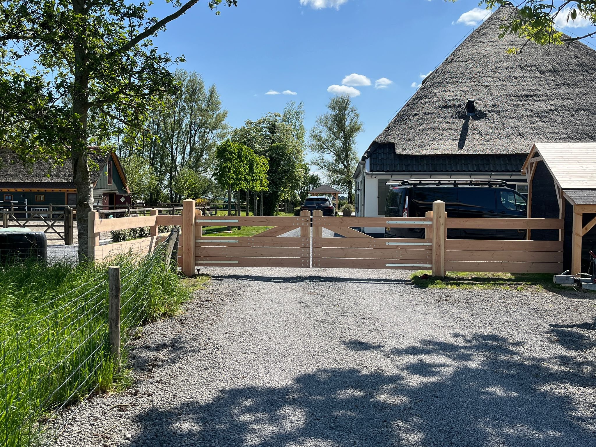 A rural scene with a gravel pathway leading to a white farmhouse with a thatched roof, surrounded by trees and greenery, with blue sky and some clouds above.