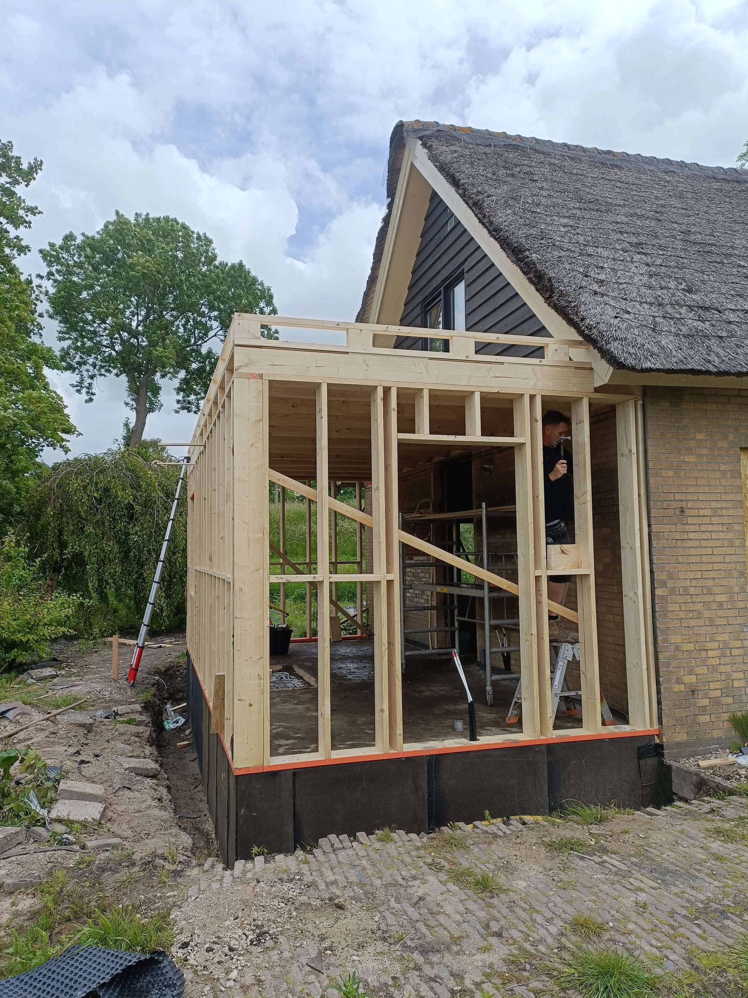 A house under construction with a wooden frame extension being built on the front of it. The main house has a steeply pitched roof with thatch and a black exterior. A person is working inside the wooden framework.