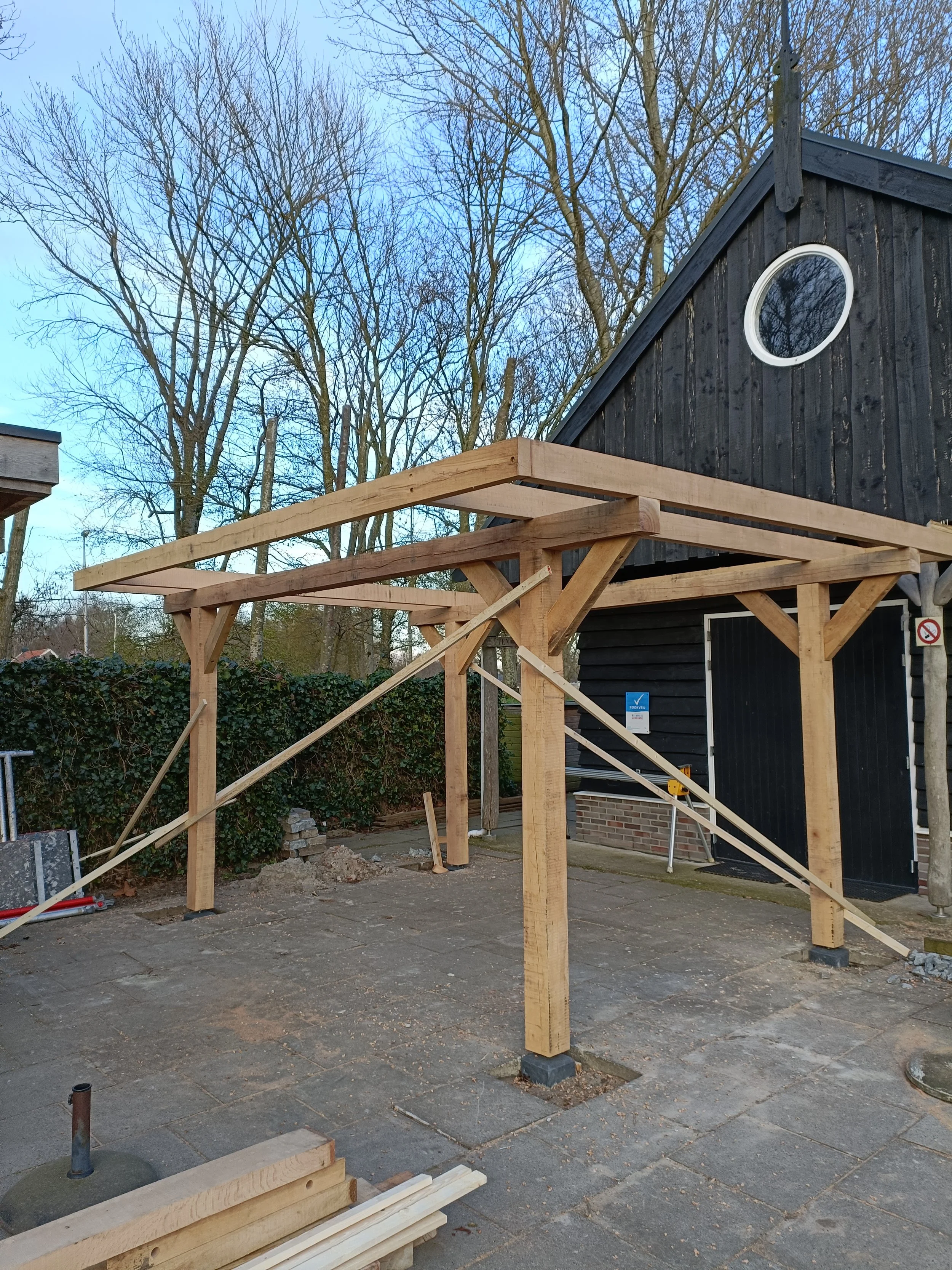Wooden construction frame in an outdoor space, with a black house in the background and leafless trees in the surrounding area.