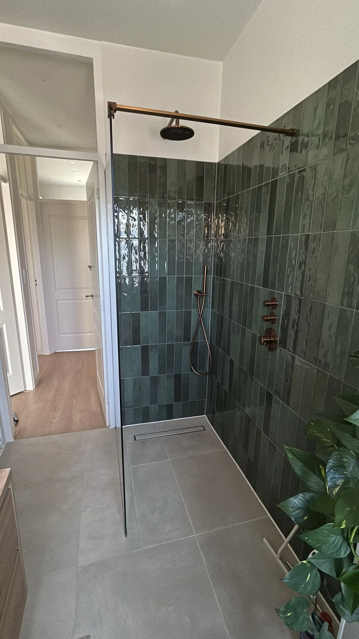 Modern walk-in shower with green tile walls, black showerhead, and brass fixtures, with a small plant visible on the right side.