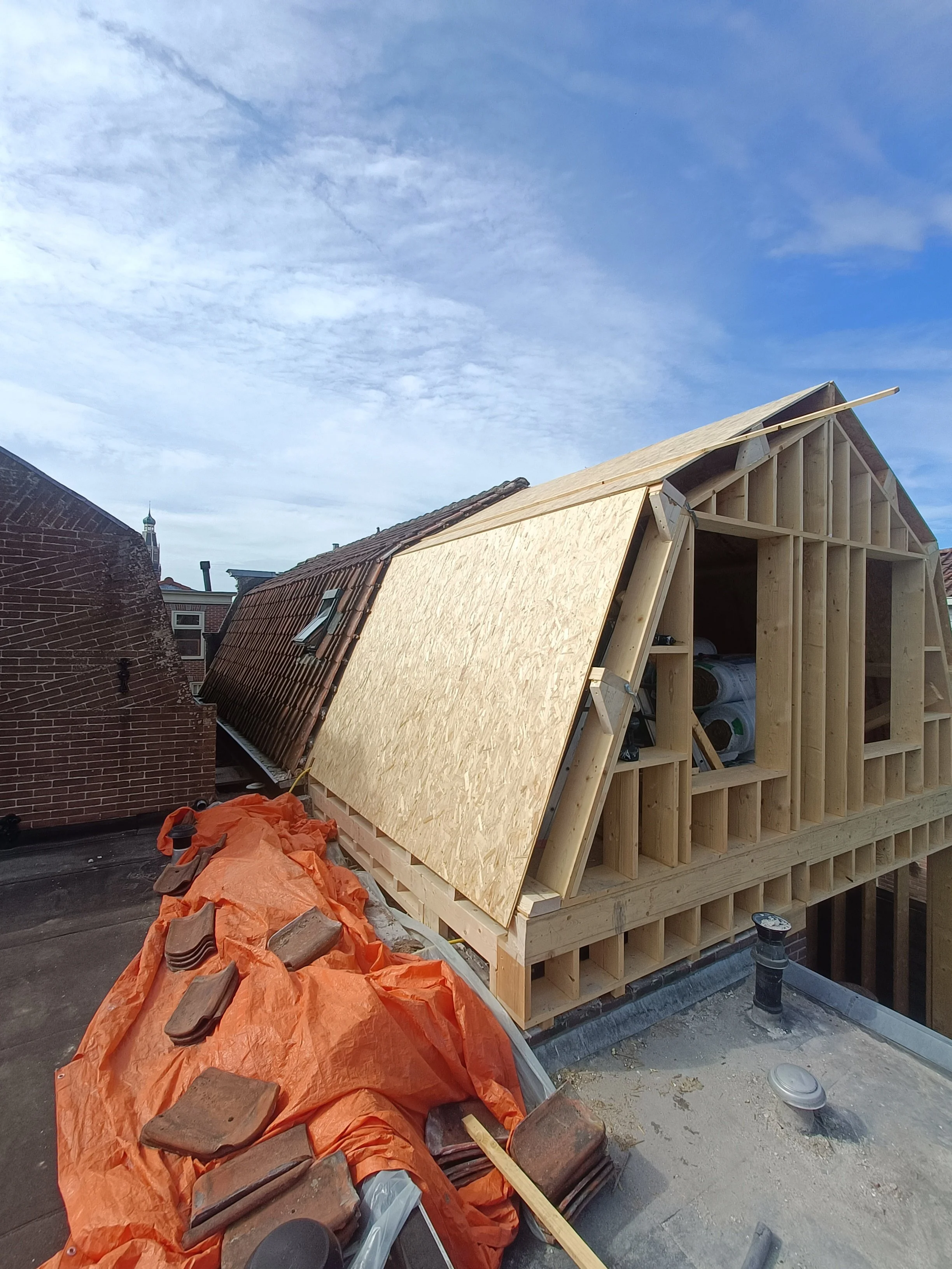 Construction site showing a partially built wooden structure with framing and a sloped roof, surrounded by construction materials and debris, under a partly cloudy sky.