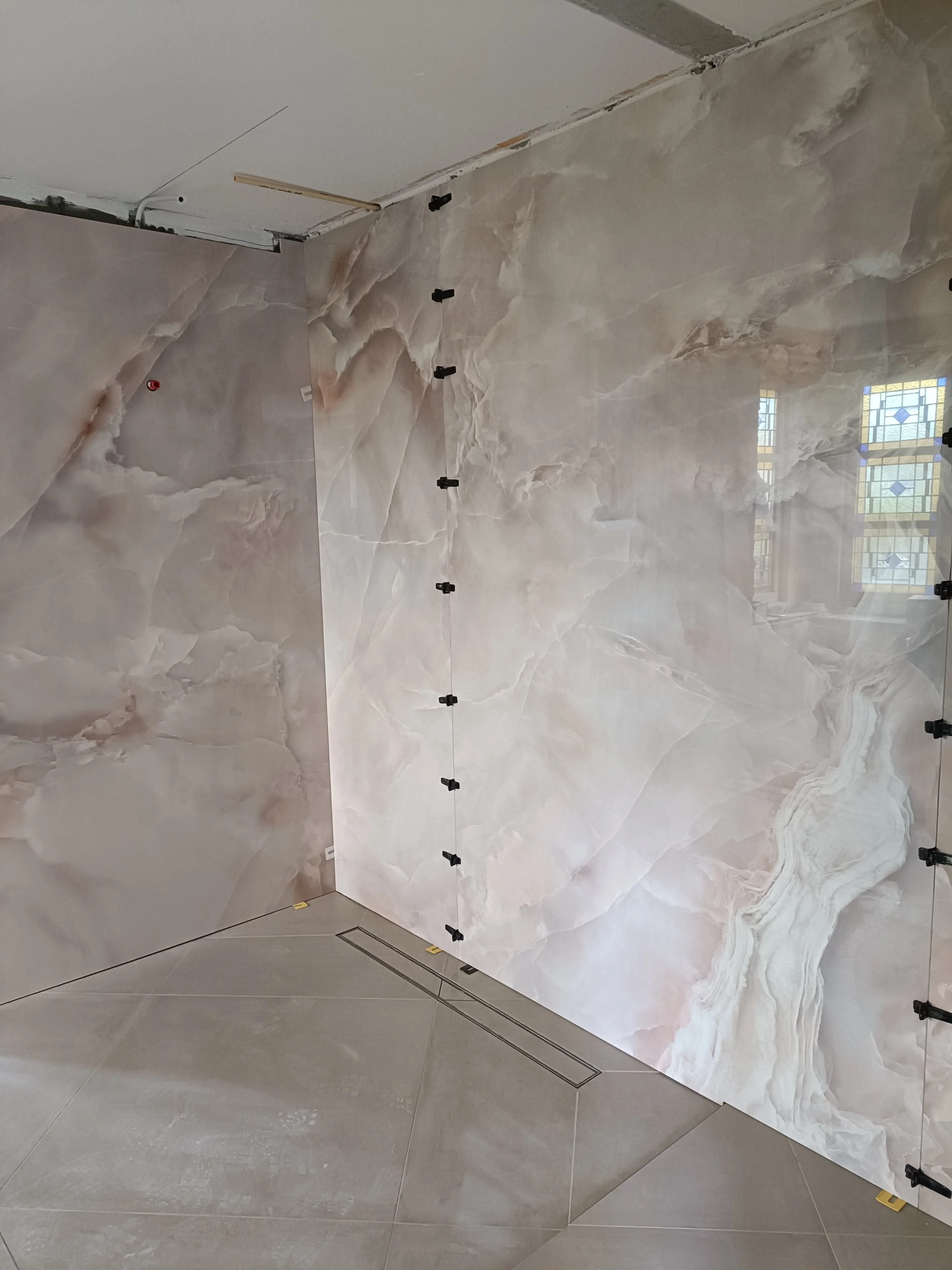 A wall with large marble tiles in progress of installation, with black clips holding the tiles in place, reflection of window panes on the glossy surface.