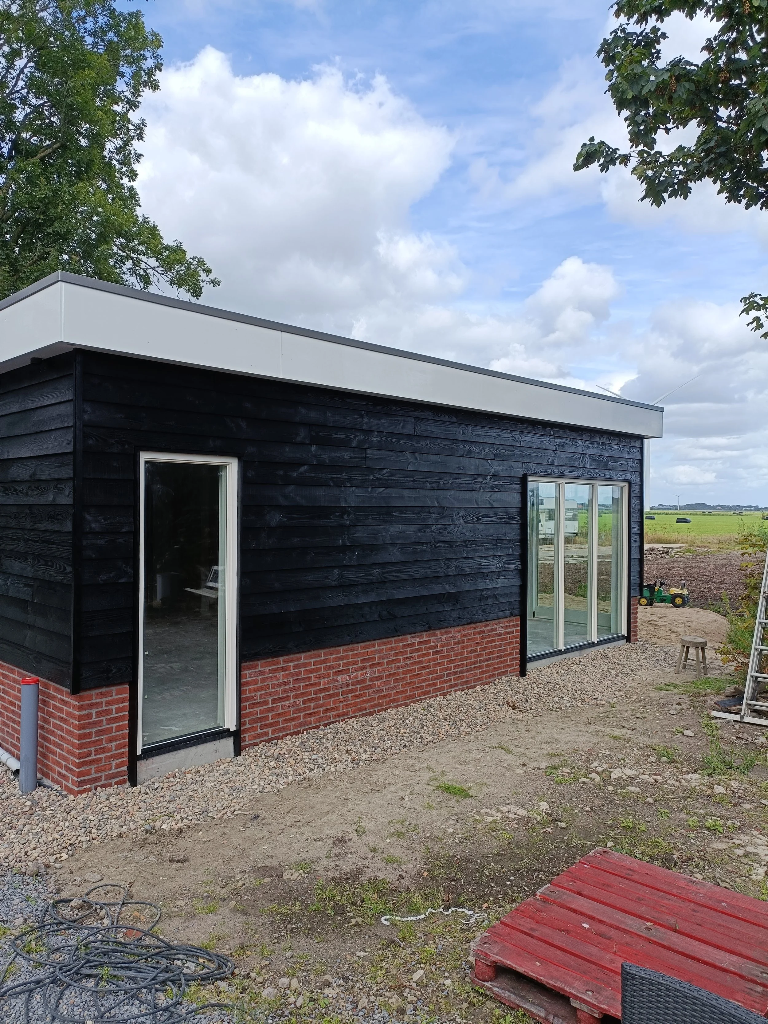 A modern house with black wooden siding and red brick foundation, large glass sliding doors, and a flat roof, situated in a rural area with open fields and a partly cloudy sky