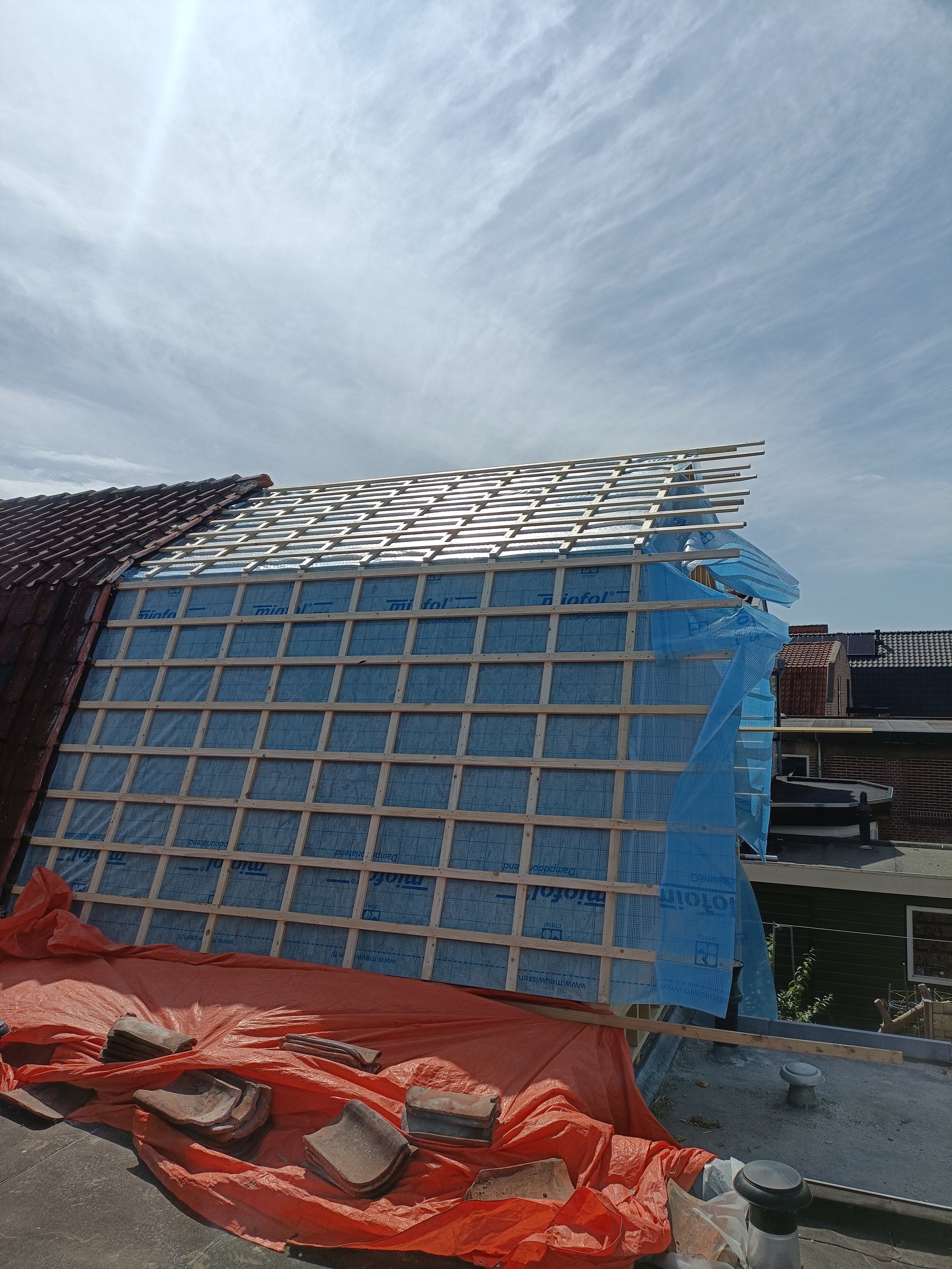 Partial view of a building under construction with roof framing and tiles, blue construction wrap, and construction materials on a red tarp in the foreground, under a partly cloudy sky.