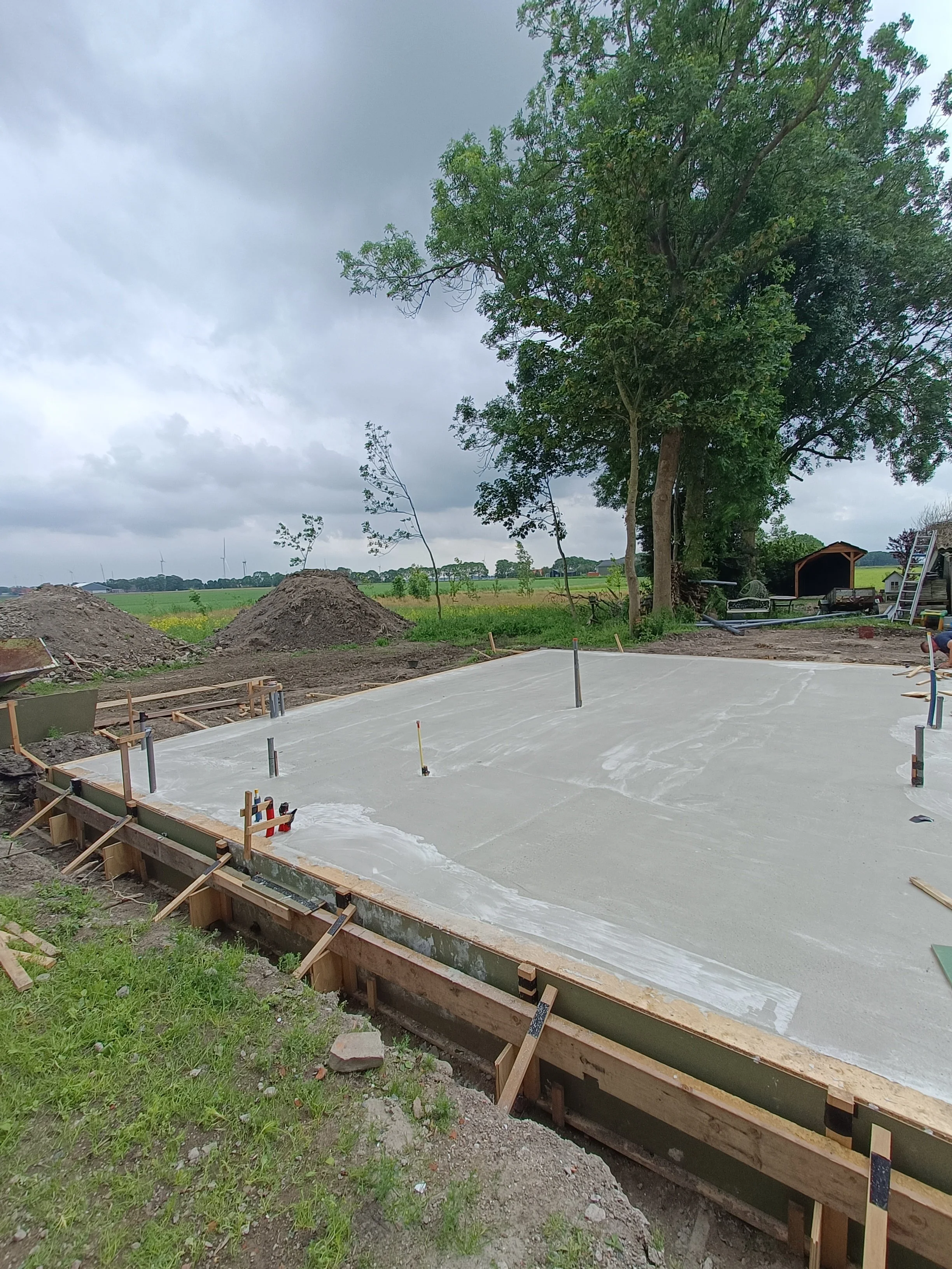 Freshly poured concrete slab on a construction site with wooden formwork, under a cloudy sky. Trees and wind turbines are visible in the distance.